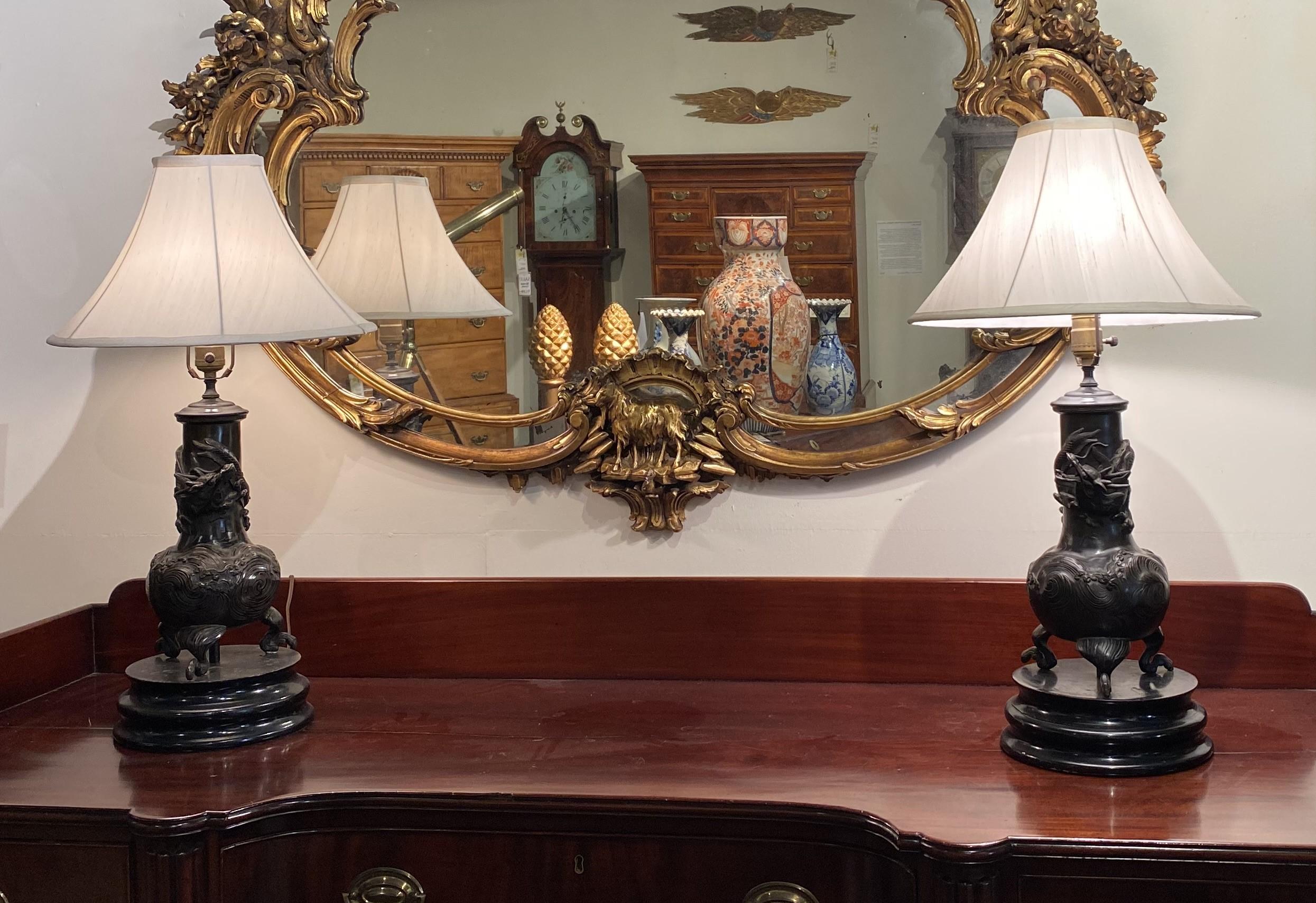 A fine pair of patinated bronze table lamps with dragon motif, on carved wooden felted bases and white flared shades, in very good working condition, with some minor surface scuffs, and light wear commensurate with age and use. The lamps are
