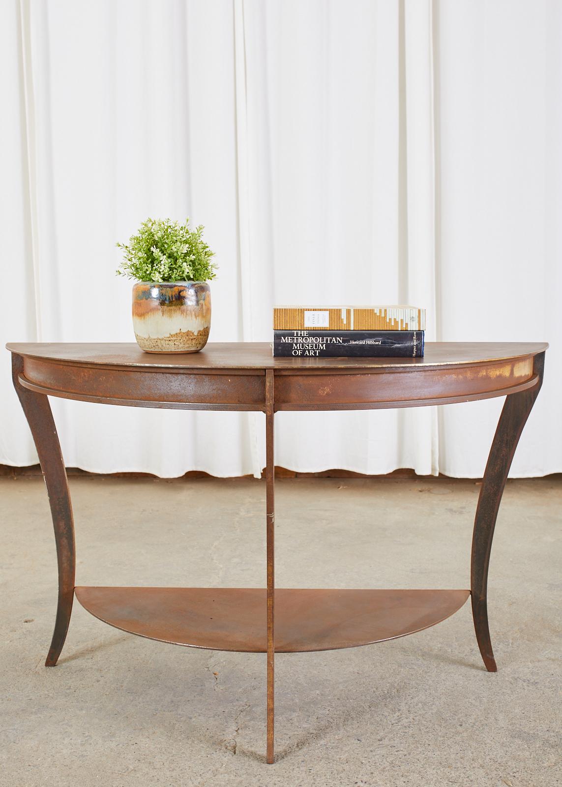 Dramatic pair of iron demi-lune console tables featuring a patinated metal finish. The elegant two-tier design has gracefully curved klismos style saber legs conjoining the shelves. The rich patinated finish could be painted, polished, or left as is