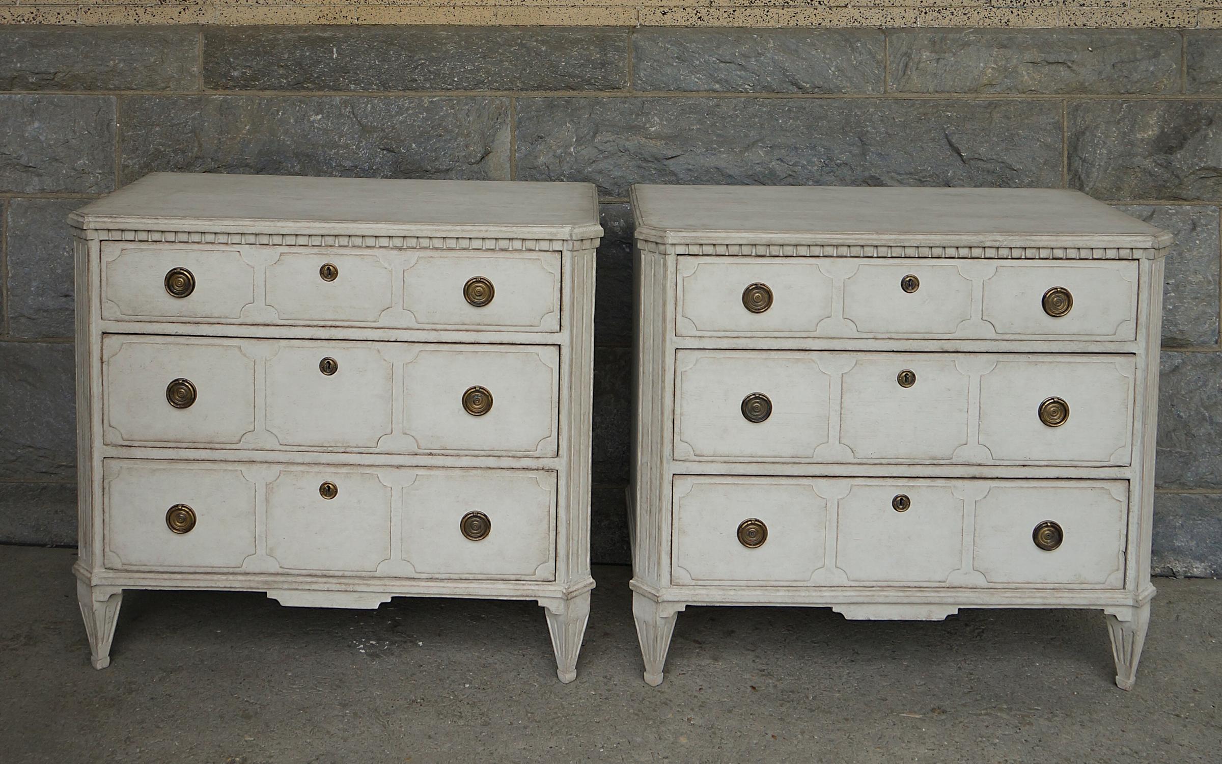 Pair of neoclassical chests of drawers in white paint, Sweden circa 1860. Each of the three graduated drawers has three raised panels and brass pulls. Dentil molding under the shaped tops and canted corners terminating in tapering square feet. All