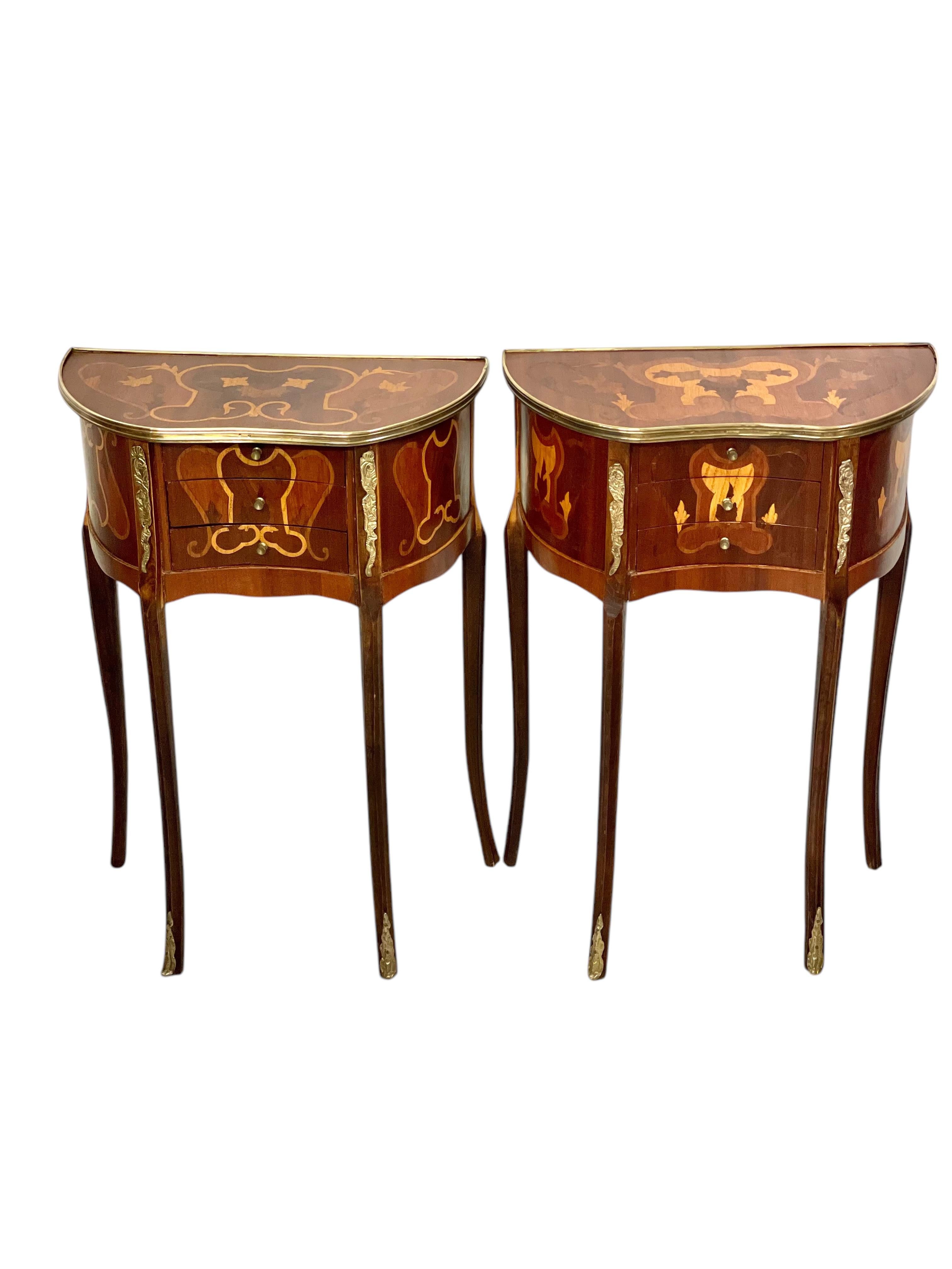 This pretty petite pair of Louis XV-style nightstands feature marquetry work, showcasing an arrangement of floral motifs and arabesques across the tops and drawers. Each console rests on four gracefully curved legs adorned with gilded brass sabots,