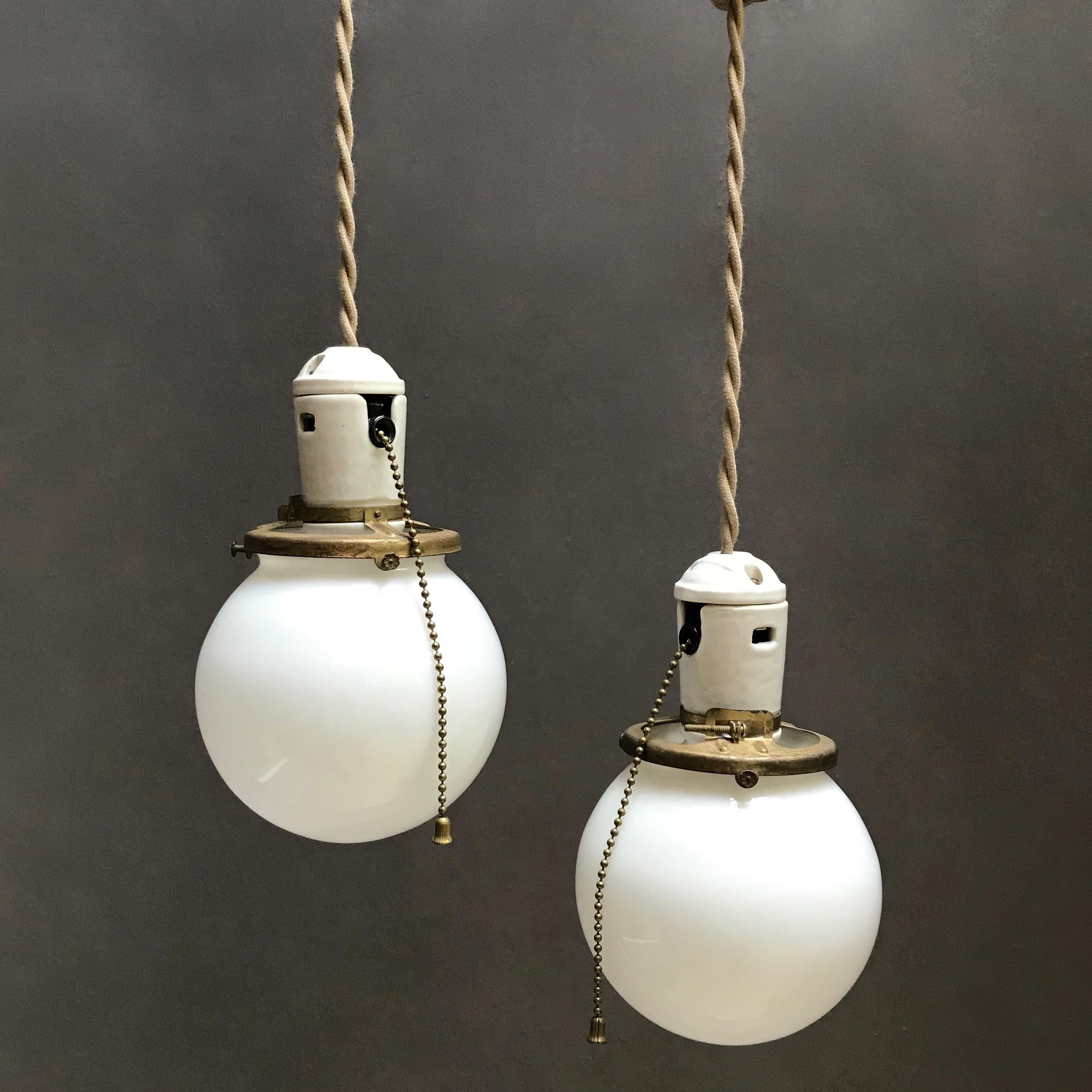 Pair of petite, Industrial, pendant lights feature milk glass globes with porcelain and brass, pull chain fitters are newly wired with beige, braided cloth cord to hang 45 inches. The lamps can accept up to 100 watt bulbs each. Listed price is for