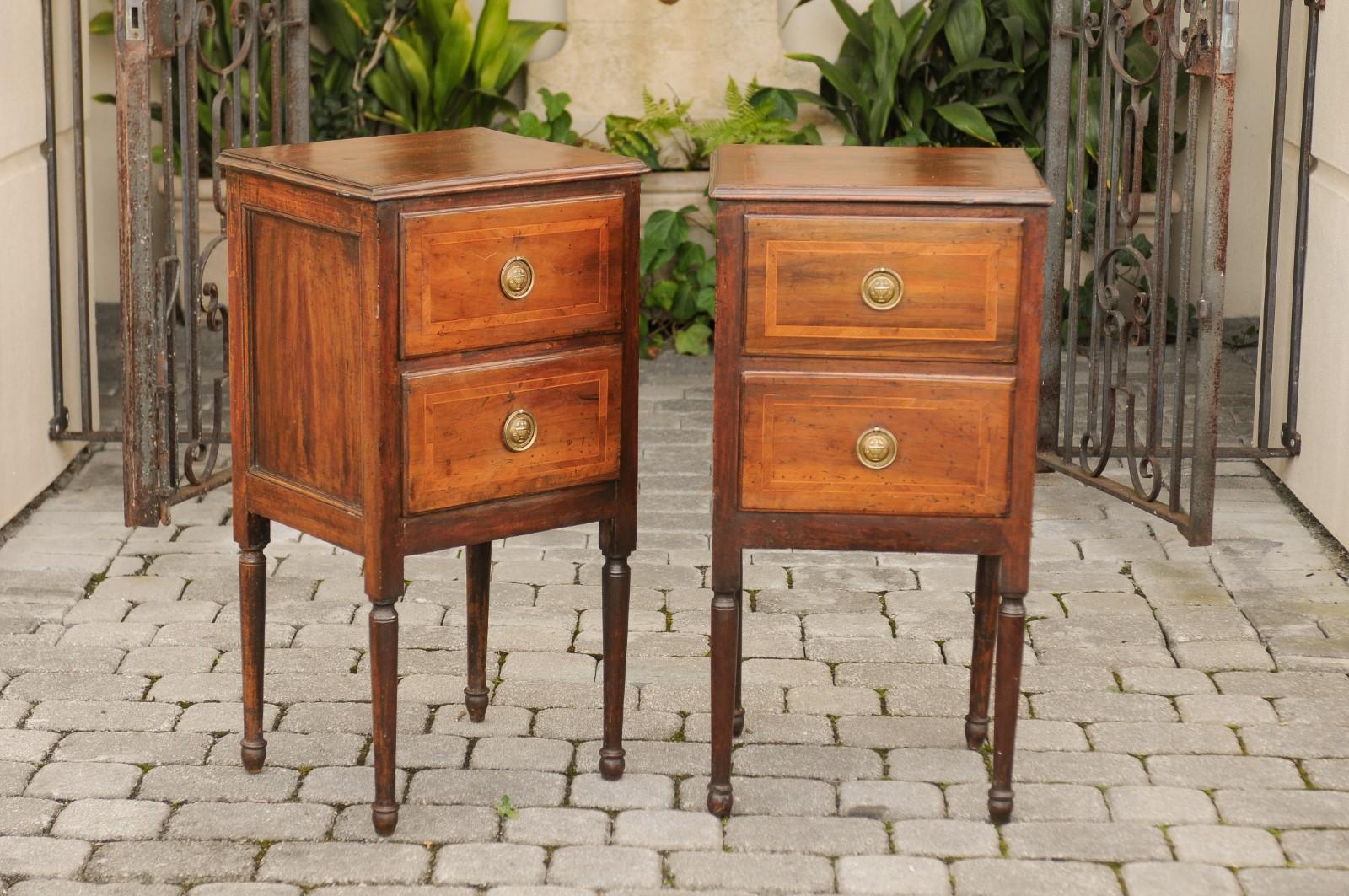 A pair of petite Italian walnut two-drawer commodes from the early 19th century with crossbanded inlay and turned legs. Each of this pair of small Italian commodes features a rectangular top, adorned with a delicate crossbanded inlay and beveled
