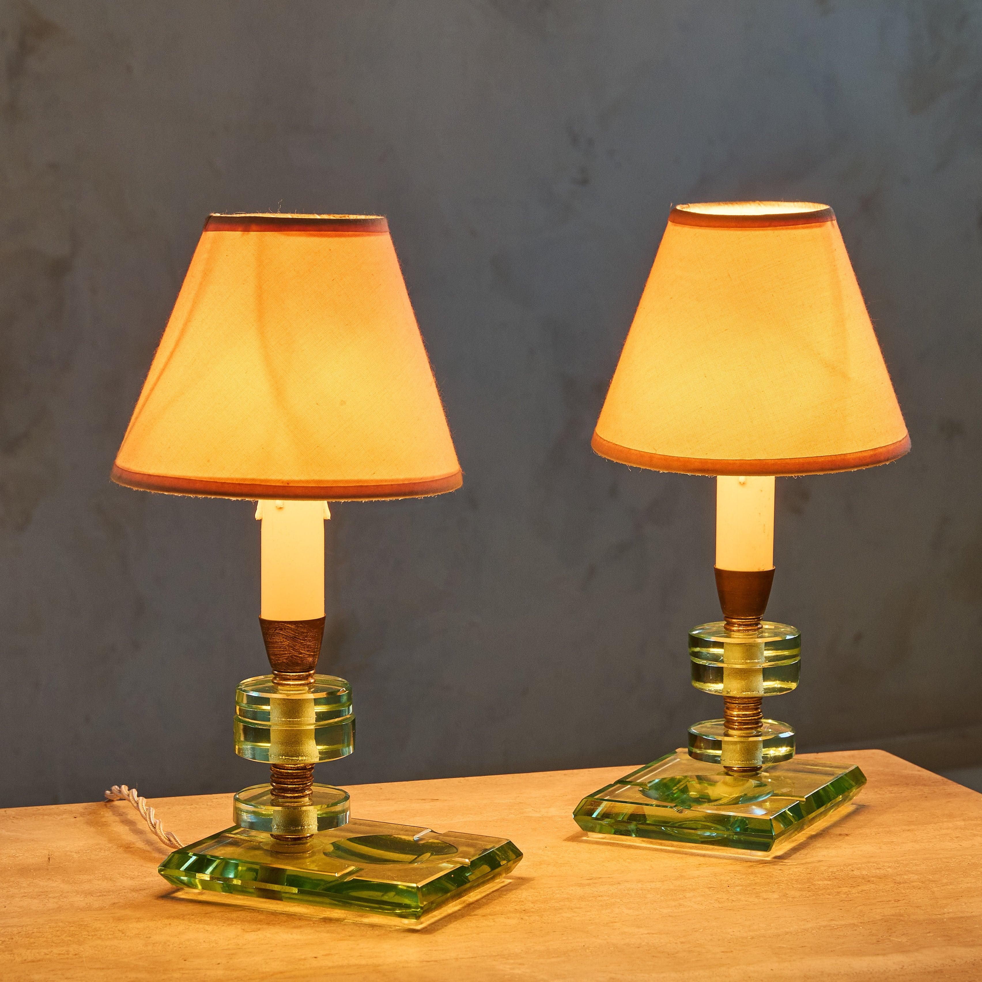 A pair of 1950s Italian candlestick-style table lamps in the manner of Fontana Arte. Crafted from thick, handblown Murano glass, the geometric bases cleverly double as catchalls. Stacked glass discs and patinated brass accents form the bodies,