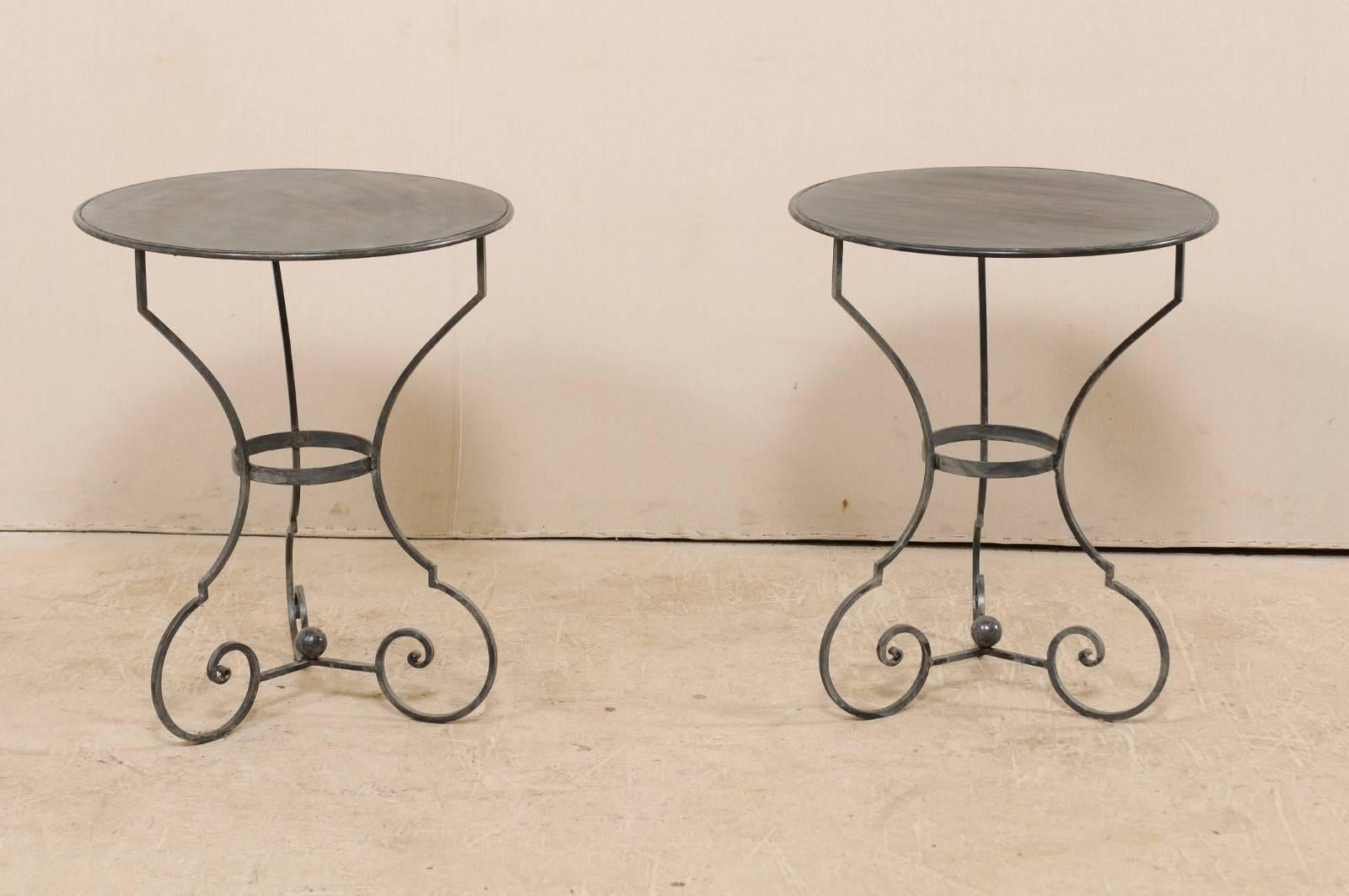 A pair of petite-sized bistro tables. This pair of round gueridon tables have been custom made and feature round metal tops, 2 feet in diameter, raised upon a steel base comprised of three fluid legs, which turn inwardly at their bottoms,