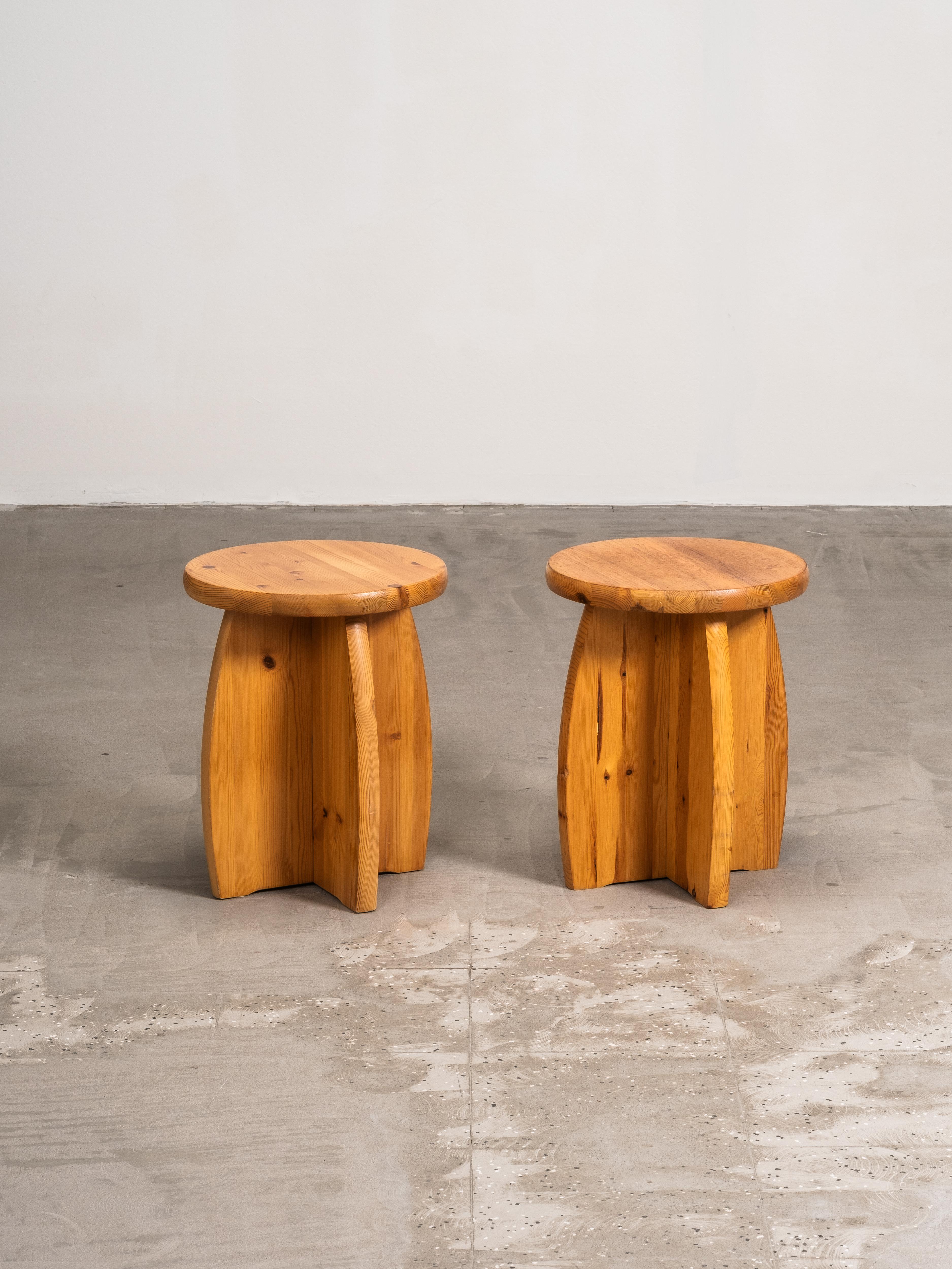 A pair of Swedish pine stools from the 1970s in a brutalist style. Each stool has a solid circular seat resting on sculptural cross-shaped legs. The design is raw and expressive, highlighting the natural grain, knots, and warm tone of solid pine.