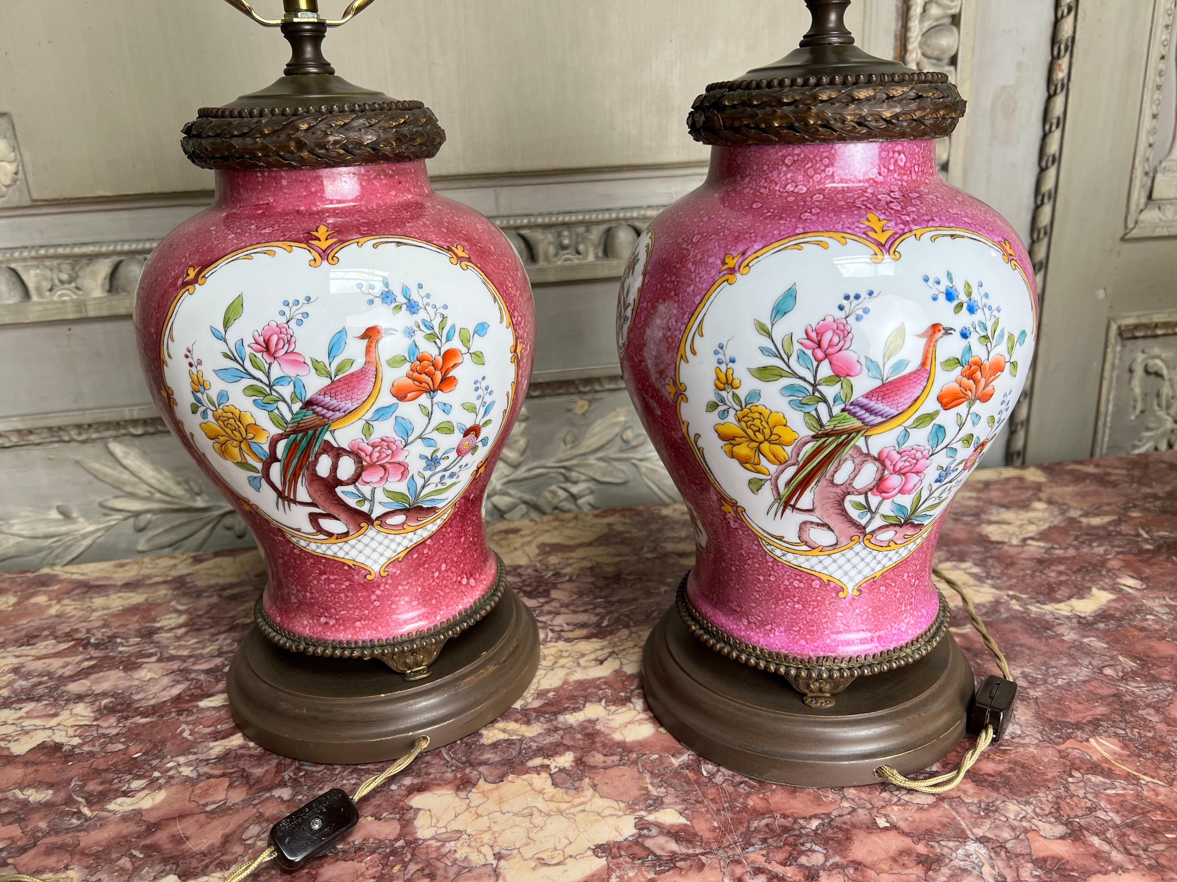 A pair of European porcelains with bronze fittings wired for lamps.  
These pink and white porcelain jars are decorated in polychrome enamel with birds and flowers.  A custom base and shade were added in the late 20th century at the time they were