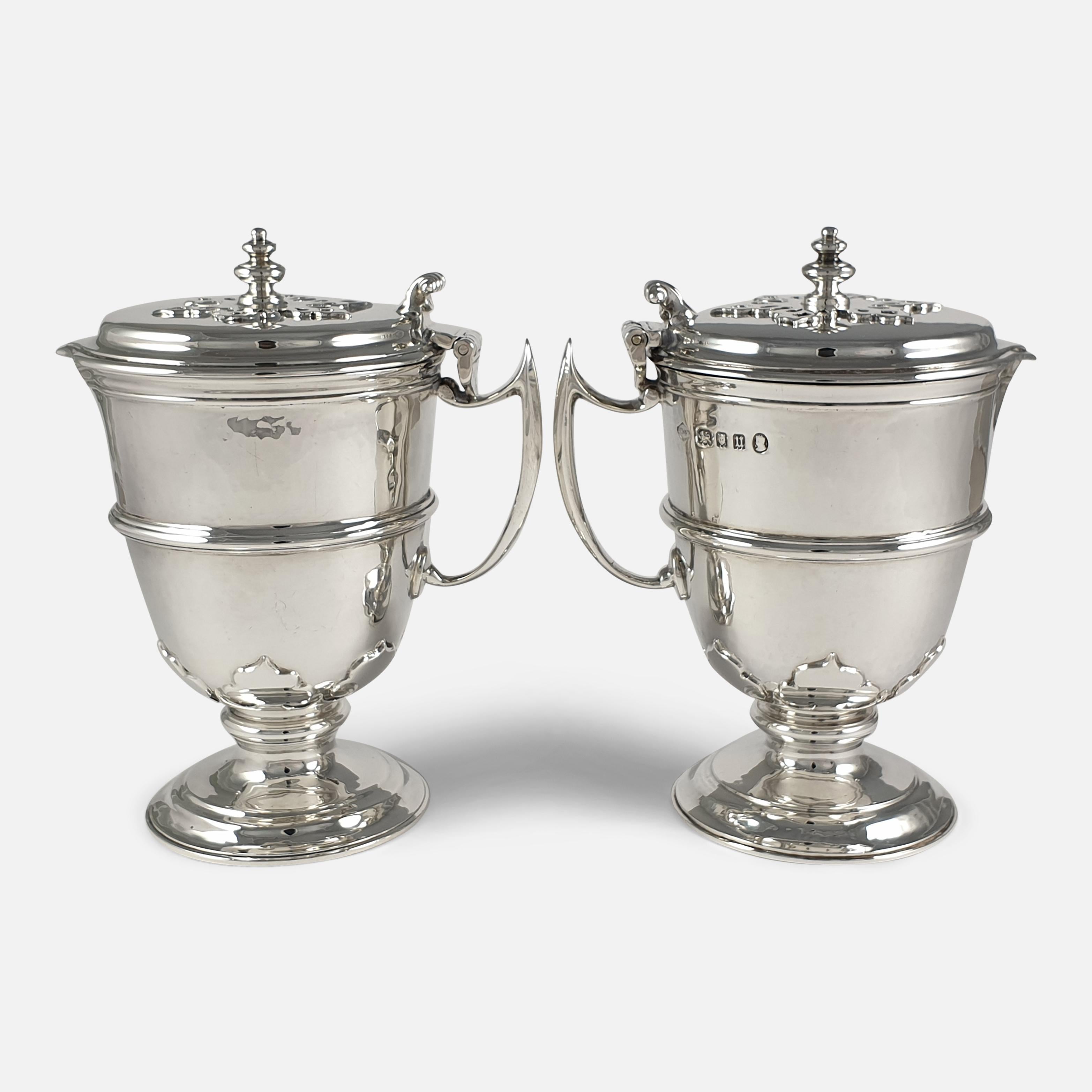 A pair of George V Arts and Crafts style sterling silver ewers. The small ewers are of planished circular form, having hinged covers with disc finial and scroll thumb-piece. The body of the jugs have a central girdle and card-cut decoration in