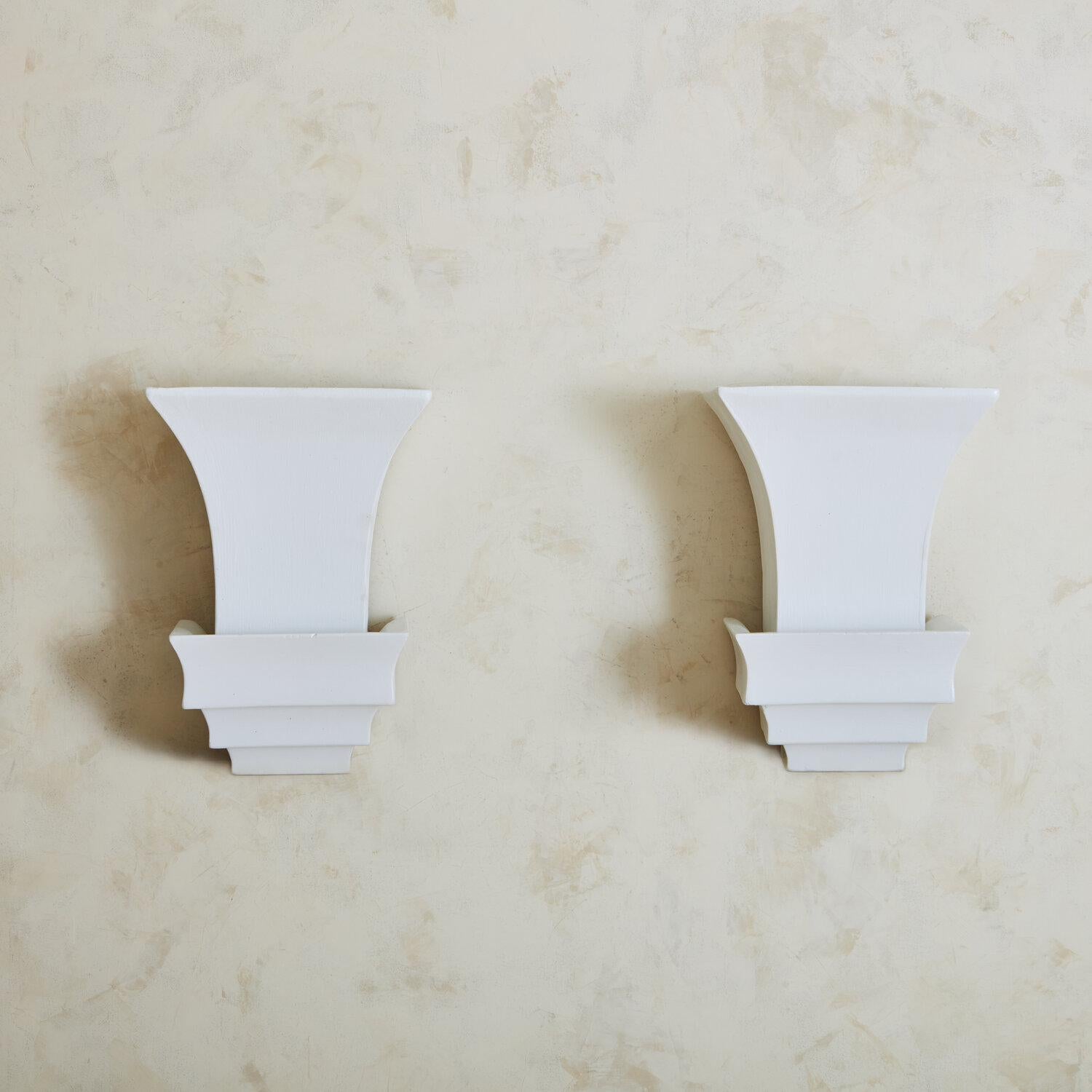 A pair of wall sconces featuring an elegant plaster three-tiered base that supports a tapered, rectangular plaster shade. The subdued white tone of these fixtures would stand out in any space and their classic shape holds a timeless appeal. 

