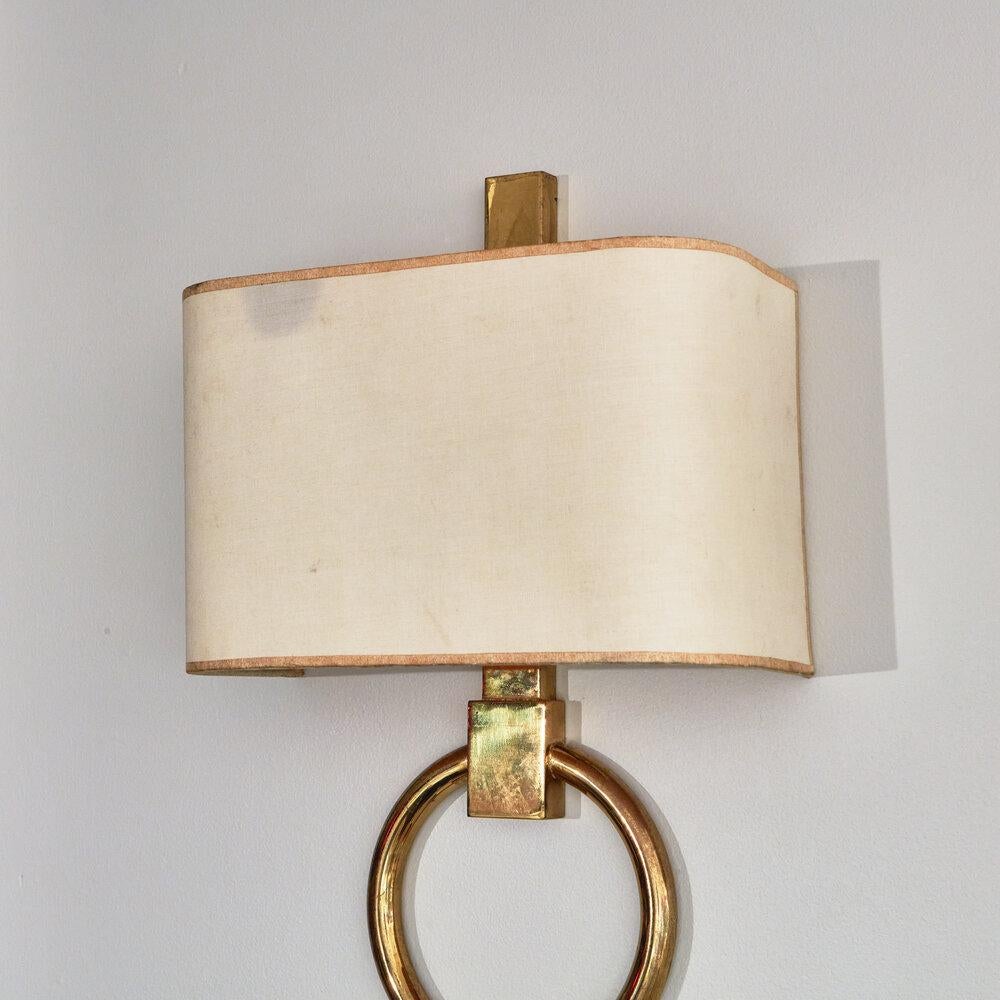A pair of handsome polished brass wall sconces featuring a suspended circular pendant and vertical frame detail that extends upwards past the rectangular linen shade. 

