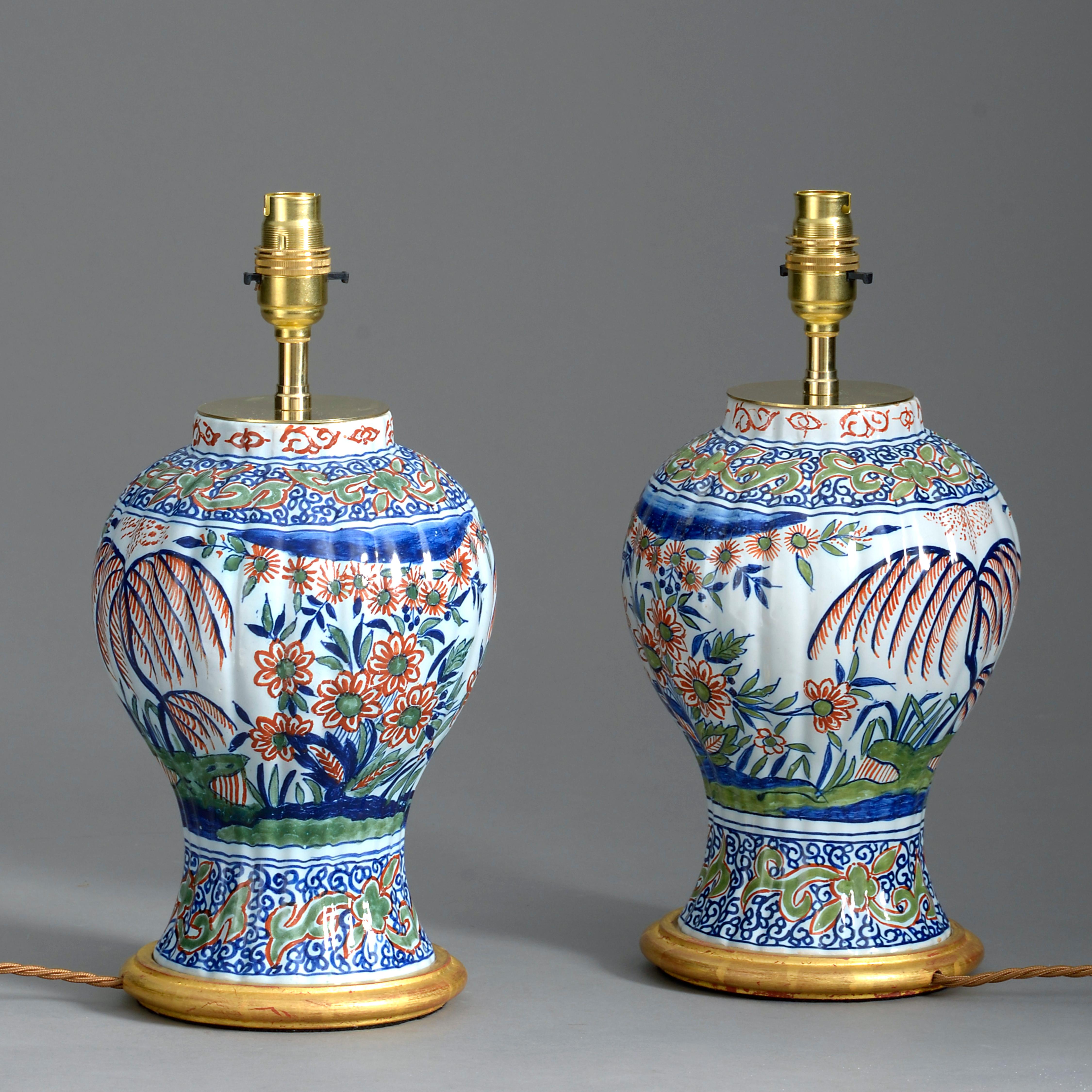 A pair of polychrome glazed delft vases of ribbed baluster form, decorated throughout with chinoiserie. Now mounted on turned water gilded bases as table lamps.

Height dimensions refer to height of pottery vases only.

Wired and tested for UK