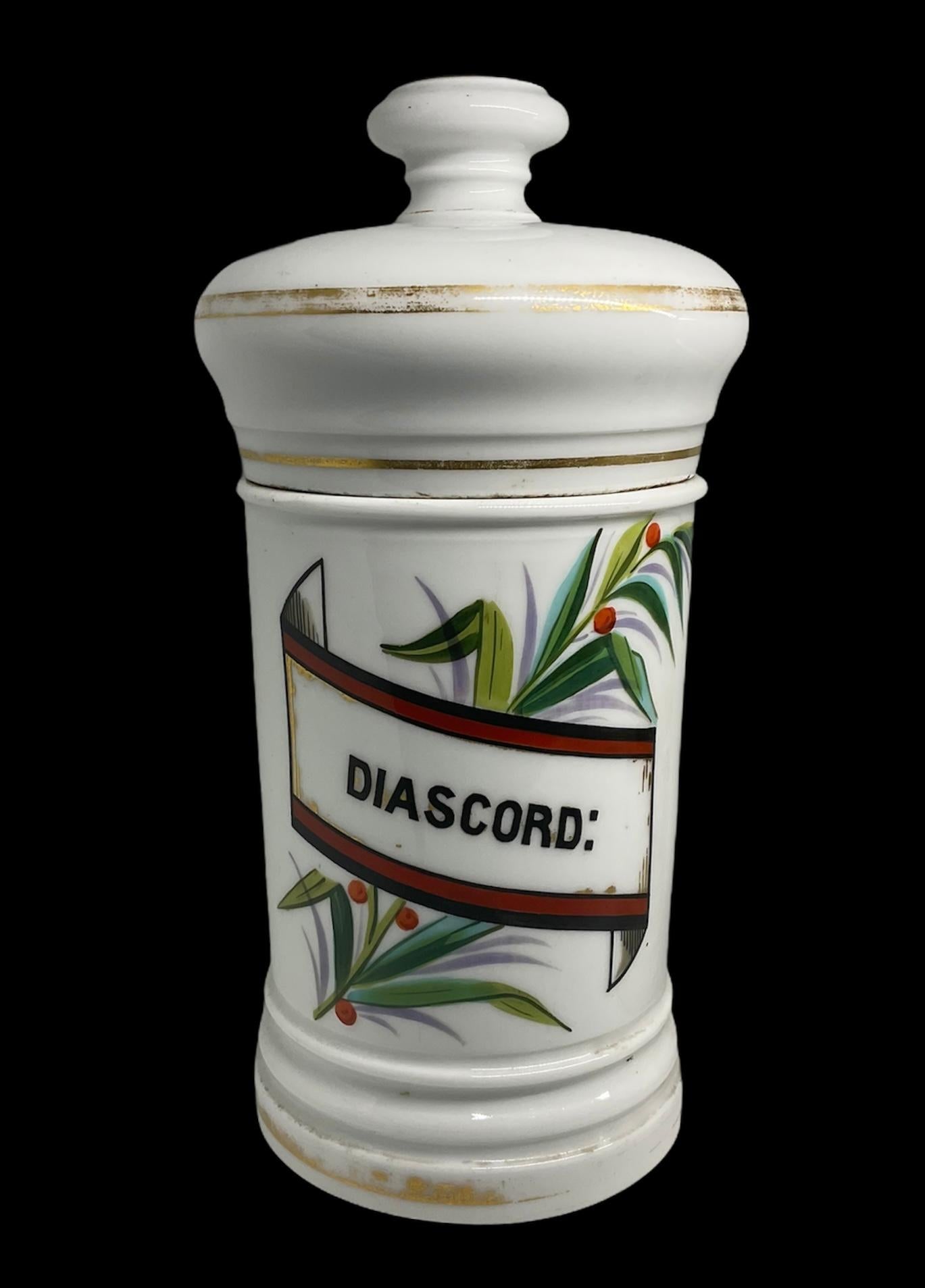 This a pair of wide cylindrical white porcelain apothecary lidded jars. They are hand painted and decorated with some laurel’s branches in the front. The lids are shaped as doctoral tams with a flatted orbit finial. They are also adorned with some