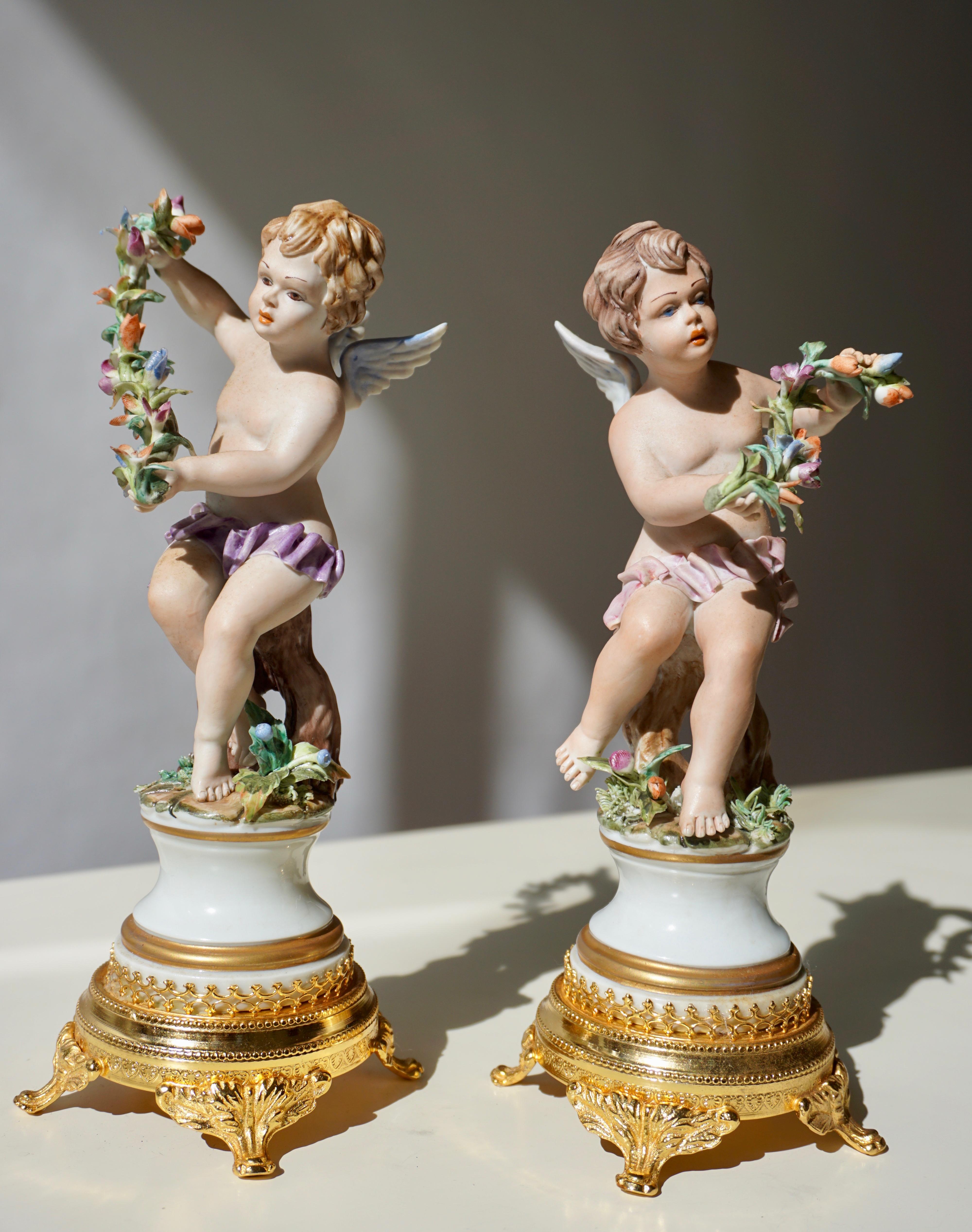 Pair of handmade polychrome porcelain sculptures depicting two winged angels, a pair of fine putti, one holding flowers, Allegory of abundance. Both porcelain figures stand on a golden circular brass base. In excellent condition, both putti figures