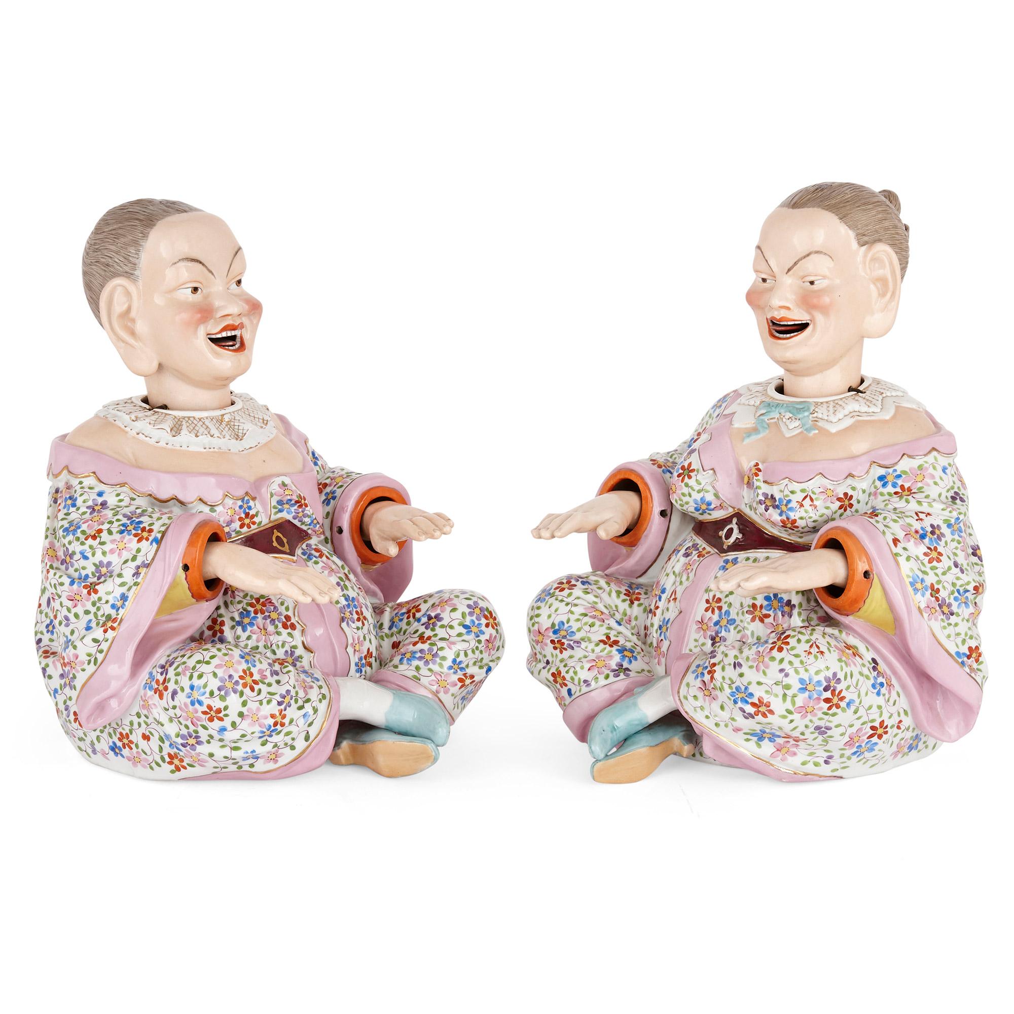 These Ernst Bohne Söhne porcelain models take the form of a Chinese couple, who sit with their legs crossed. They have large bellies and ears, and broad grins on their faces. Both figures wear parcel gilt collared shirts and loose robes, decorated