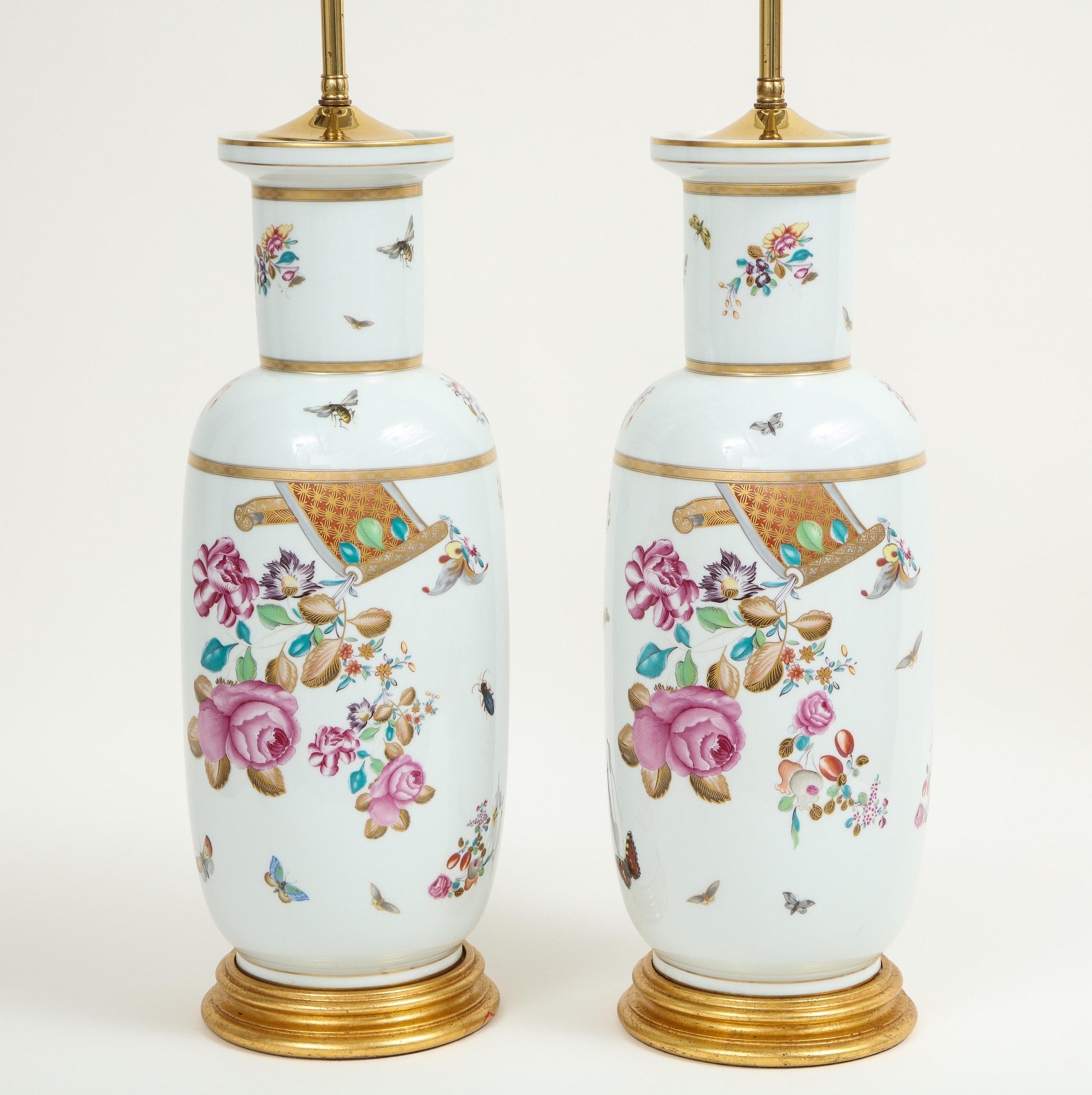Each decorated in the 18th century spirit with floral blossoms, butterflies and bees, and gilt banding, on a giltwood turned base. Fitted with brass sockets for two bulbs and adjustable rod to change lampshade height. Height to top of vase is 20