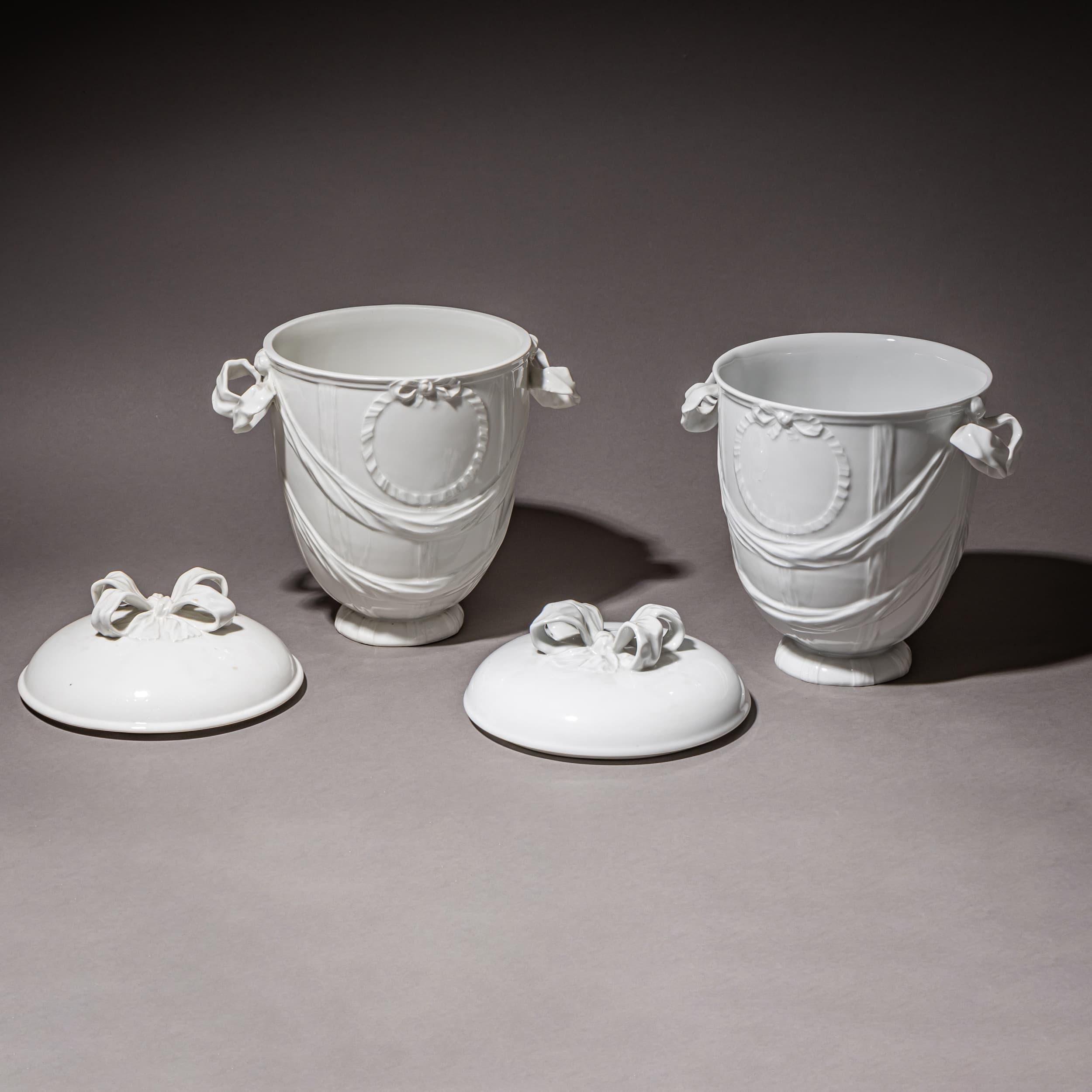 Pair of lidded urns in white-glazed porcelain by KPM Berlin, with ribbon handles and draped festoons on the body. The conical, slightly bulbous vessels feature a central cartouche. The knop is shaped as a ribbon. Each with blue underglaze sceptre