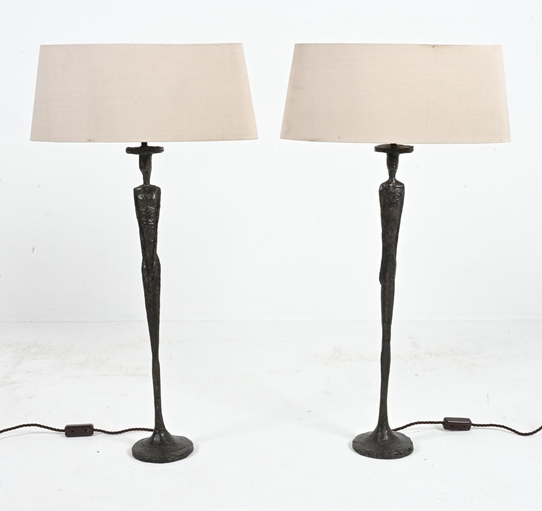 An elegant pair of figurative table lamps in tribute Alberto Giacometti. 
Patinated bronze-finished resin. 
Porta Romana, Model No. VLB/05. 
Italy, c. 2008. 
Shades included. 
Note: Dimensions provided include shades. Lamp base without shade