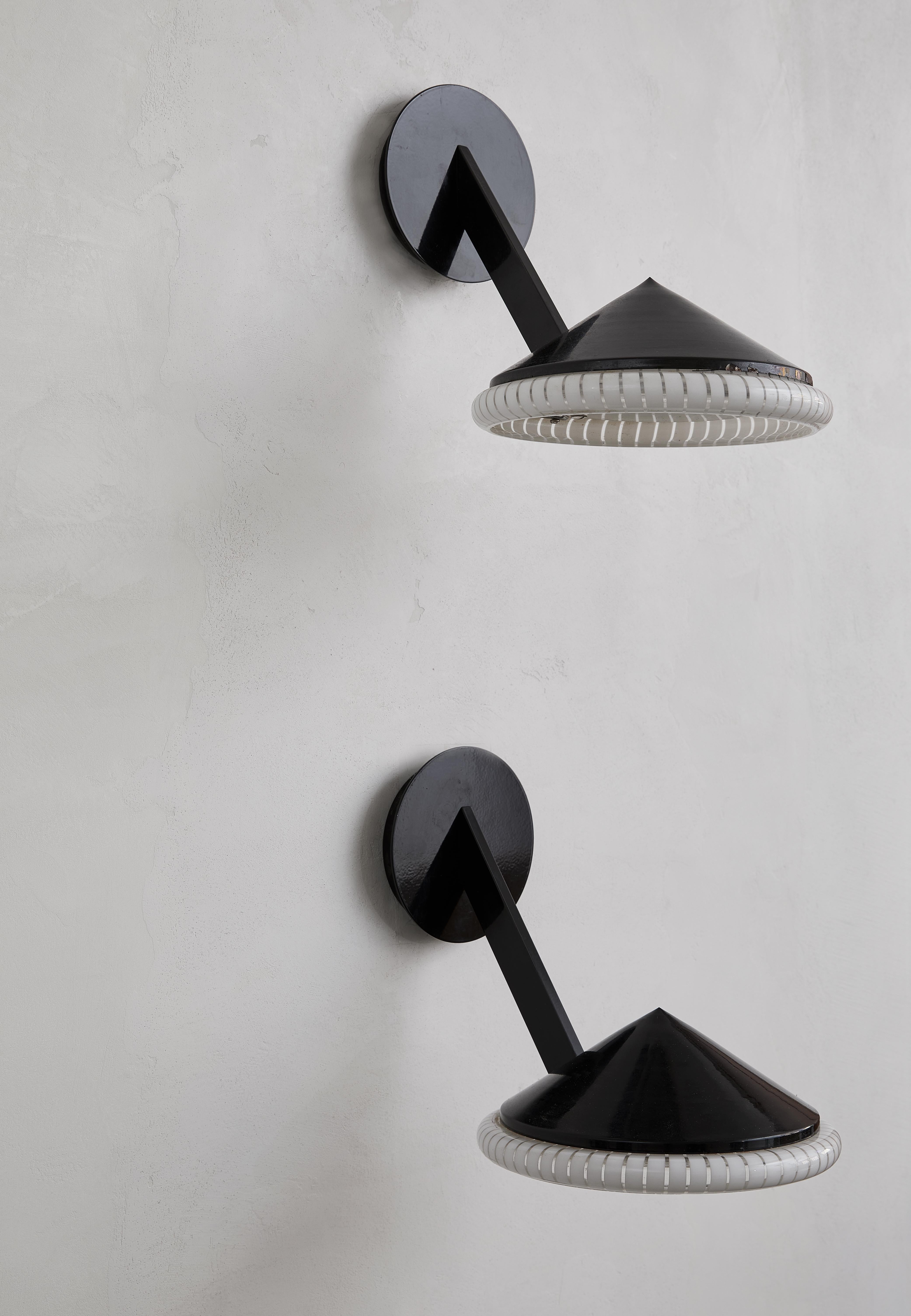 Cette paire d'appliques murales post-modernes présente un jeu audacieux de matériaux et de formes. Les cadres en métal émaillé noir créent une silhouette élégante et moderne, tandis que les diffuseurs en verre doucement frotté émettent une lueur