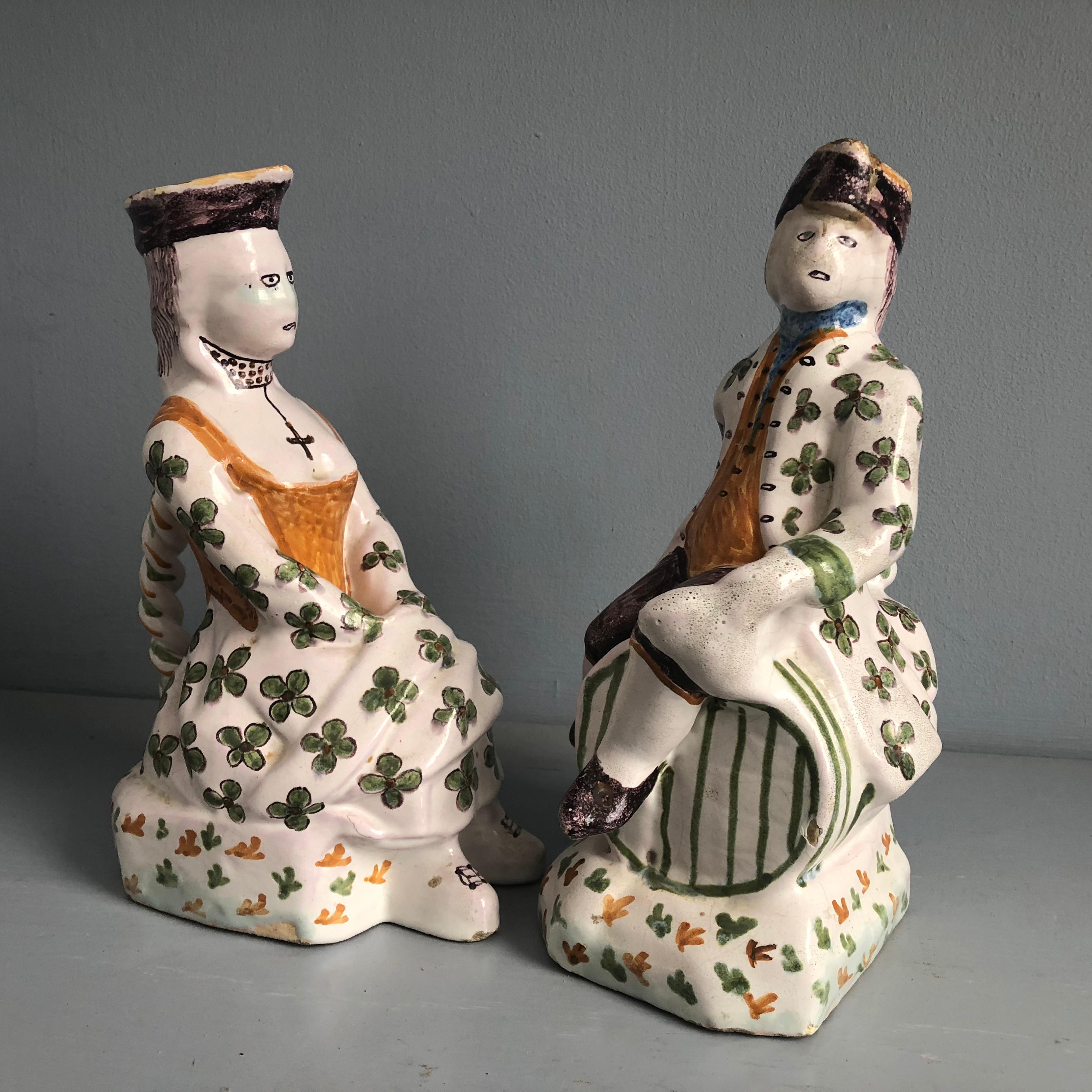 A charming pair of Faience pitchers, French Provincial late 19th century, depicting an 18th century man and a woman seated, their matching caps forming the spouts, both decorated with green clovers and other details over a white glaze. Each have a