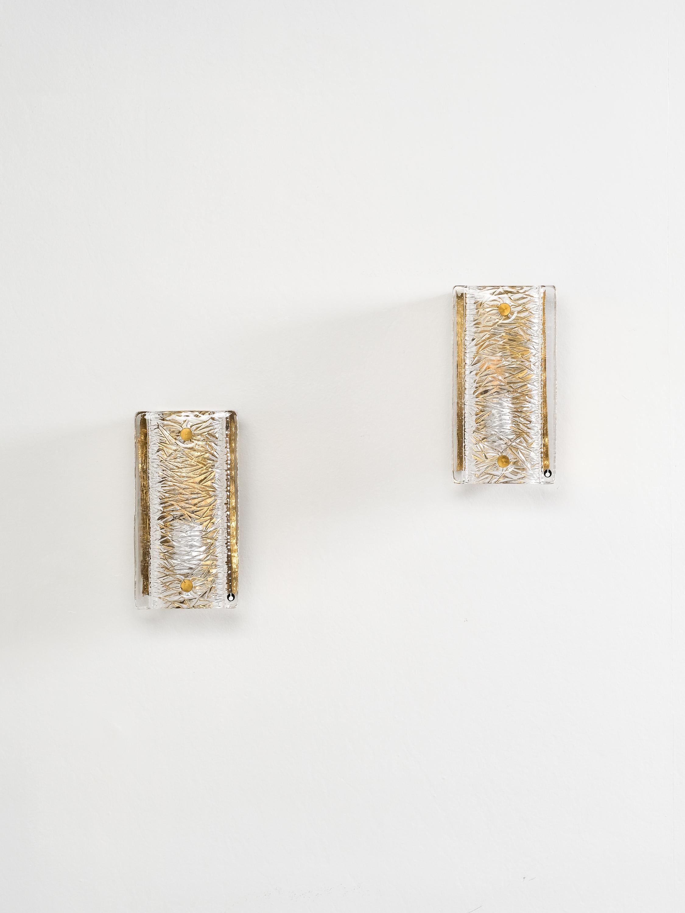 A striking pair of wall sconces by renowned Swedish glassworks Pukeberg, crafted in the 1970s. Each sconce features a polished brass backplate paired with a sculptural crystal glass shade, creating a beautiful interplay of light and texture. The