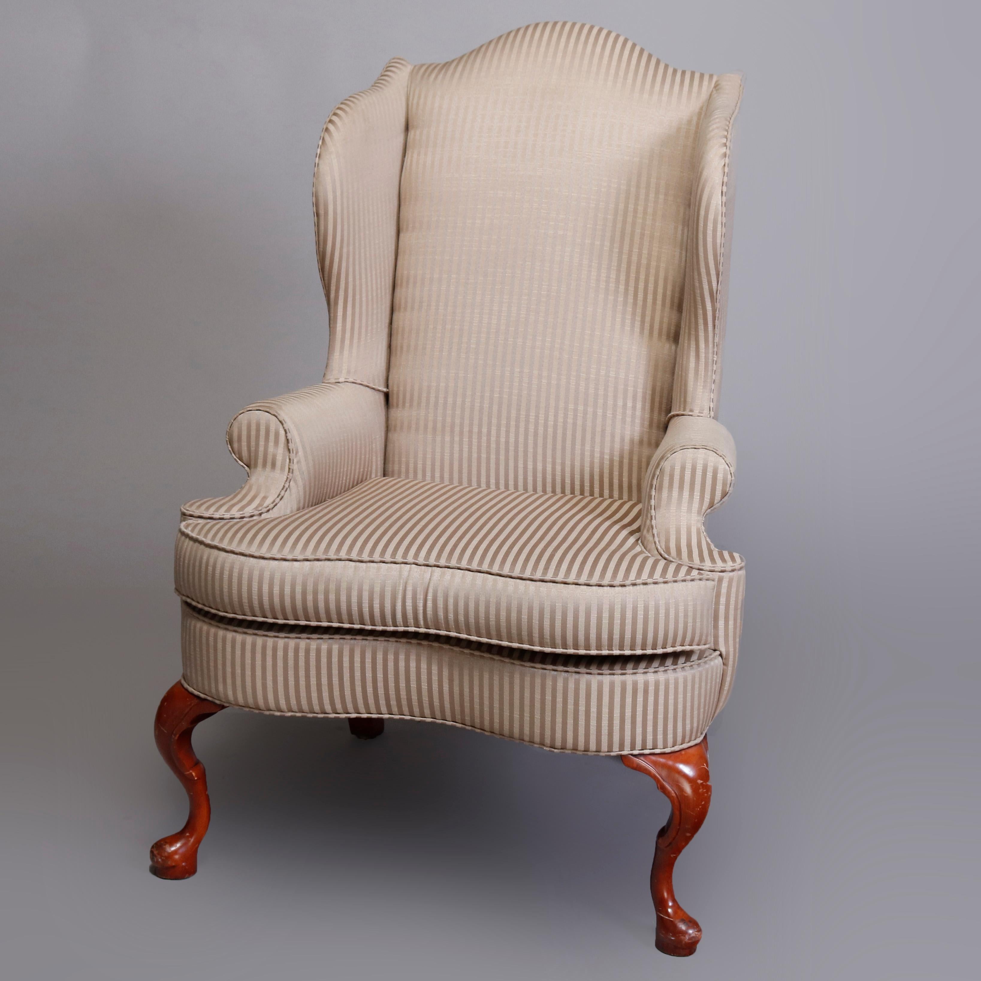 A pair of Queen Anne style fireside chairs offer wingback form upholstered with striped patterned fabric and having scrolled arms and shaped seat surmounting carved mahogany cabriole legs with pad feet, 20th century

Measures: 48