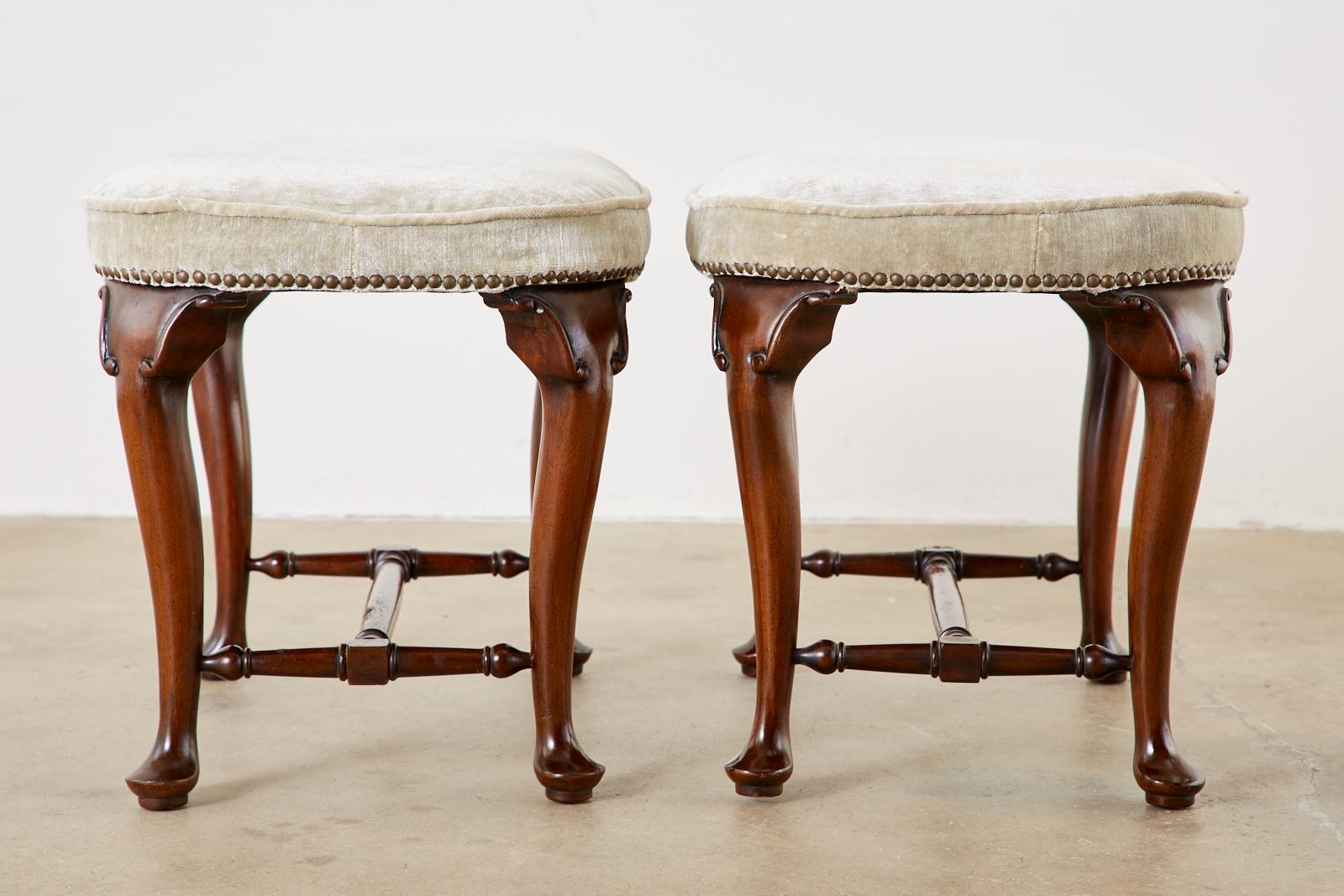 19th century matching pair of mahogany footstools made in the Queen Anne taste. Featuring plush, velvet upholstery bordered by brass tack nail heads. Supported by cabriole legs ending with pad feet conjoined by turned stretchers. From an estate in