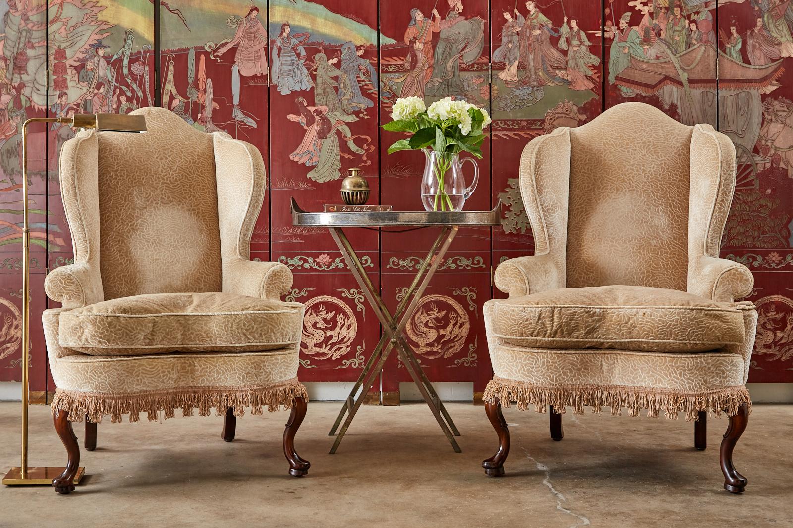 Distinctive pair of upholstered wing chairs by Dunbar. The wingbacks feature a hardwood frame with tall peaked crests and fully developed wings. The chairs are crafted in the Queen Anne taste with cabriole legs in the front ending with pad feet. The