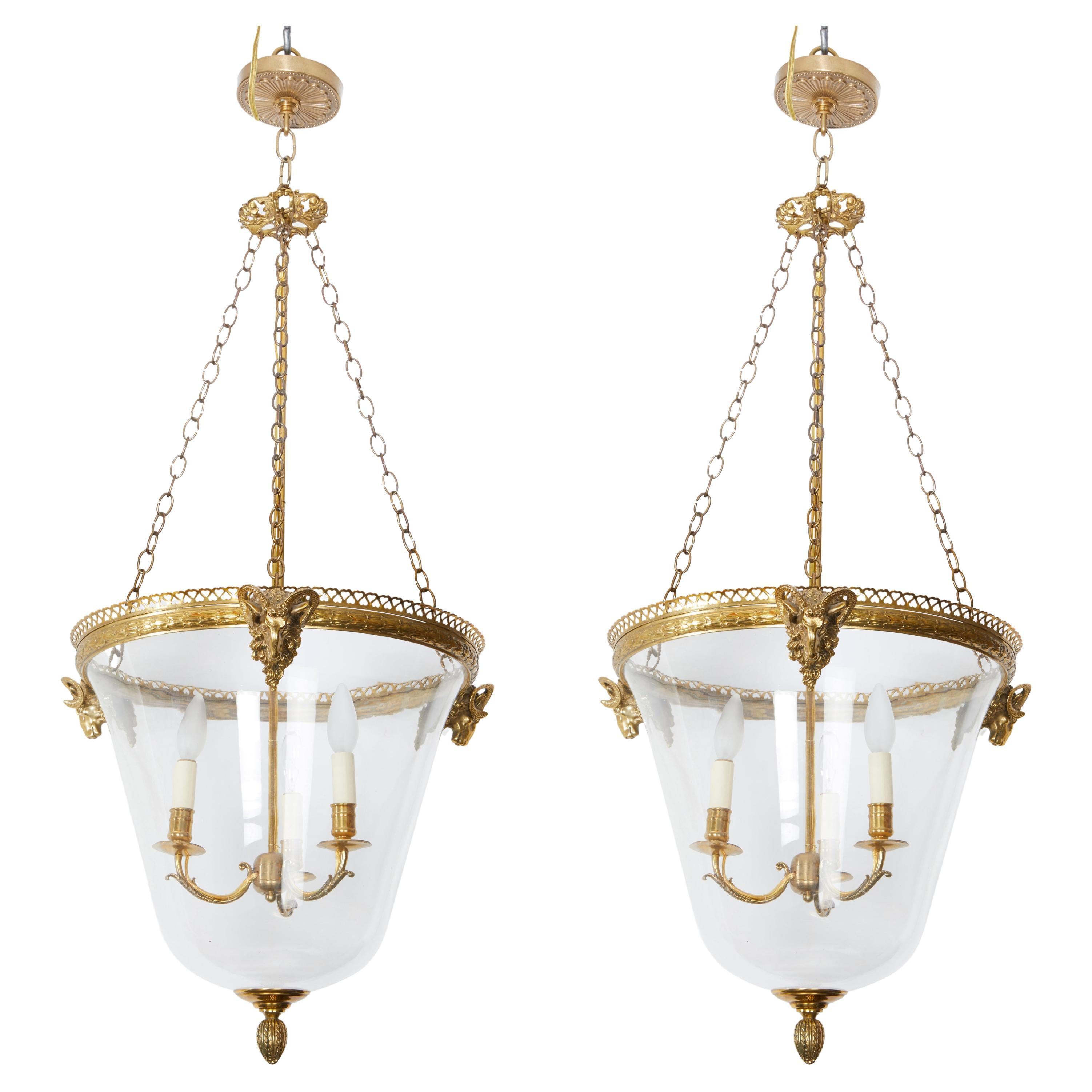A French Louie XIV style glass lantern. The bell-shape body is suspended from 3 lengths of brass chain secured to the brass rim with pierced gallery edge. Three Ram heads mount each lantern with a three light cluster suspended from a canopy while