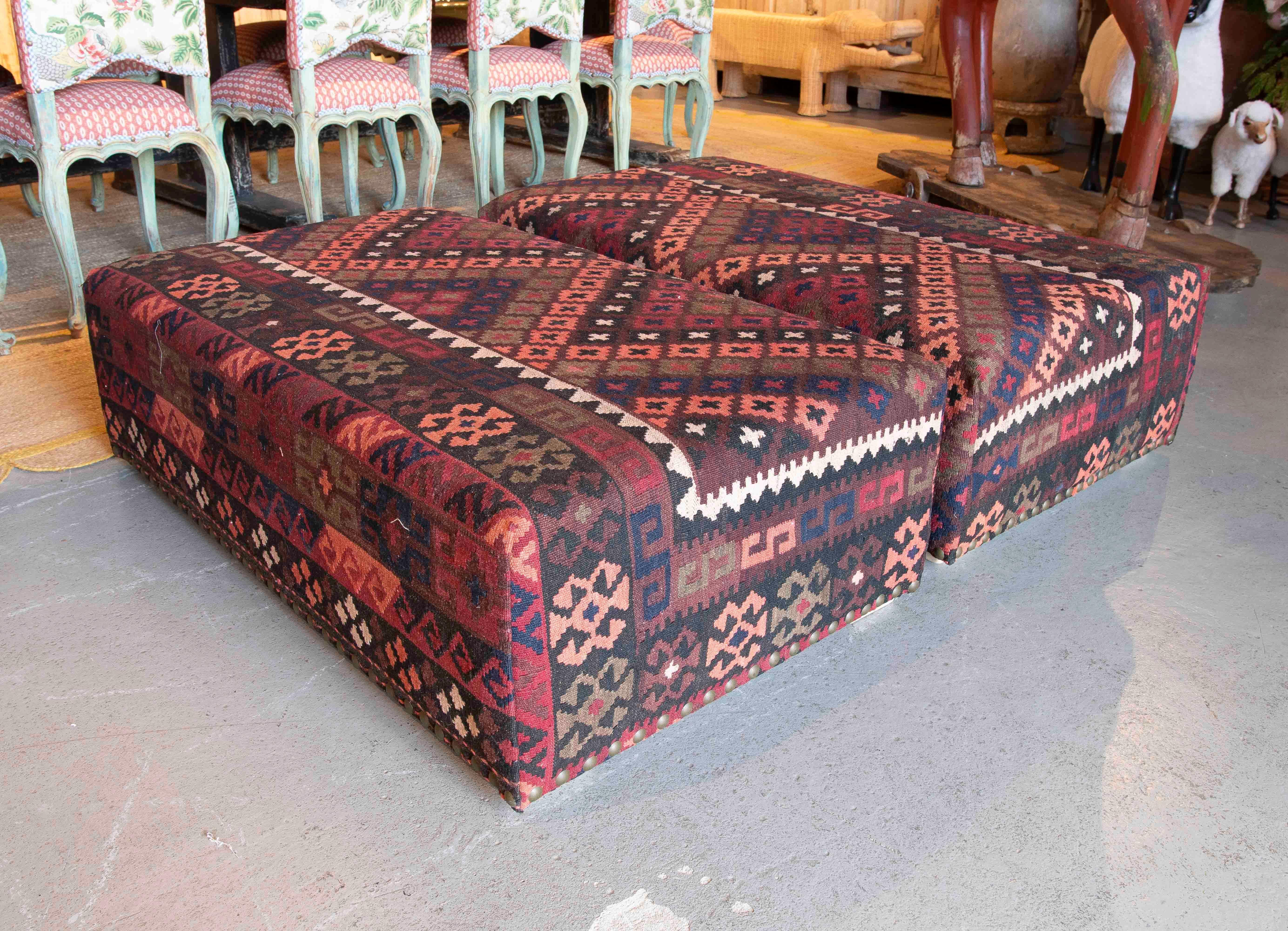 This elegant pair of rectangular poufs is upholstered with ethnic-inspired kilim fabric, highlighting vibrant geometric patterns in warm tones such as red, black and beige. The sturdiness of their structure and unique design make them functional