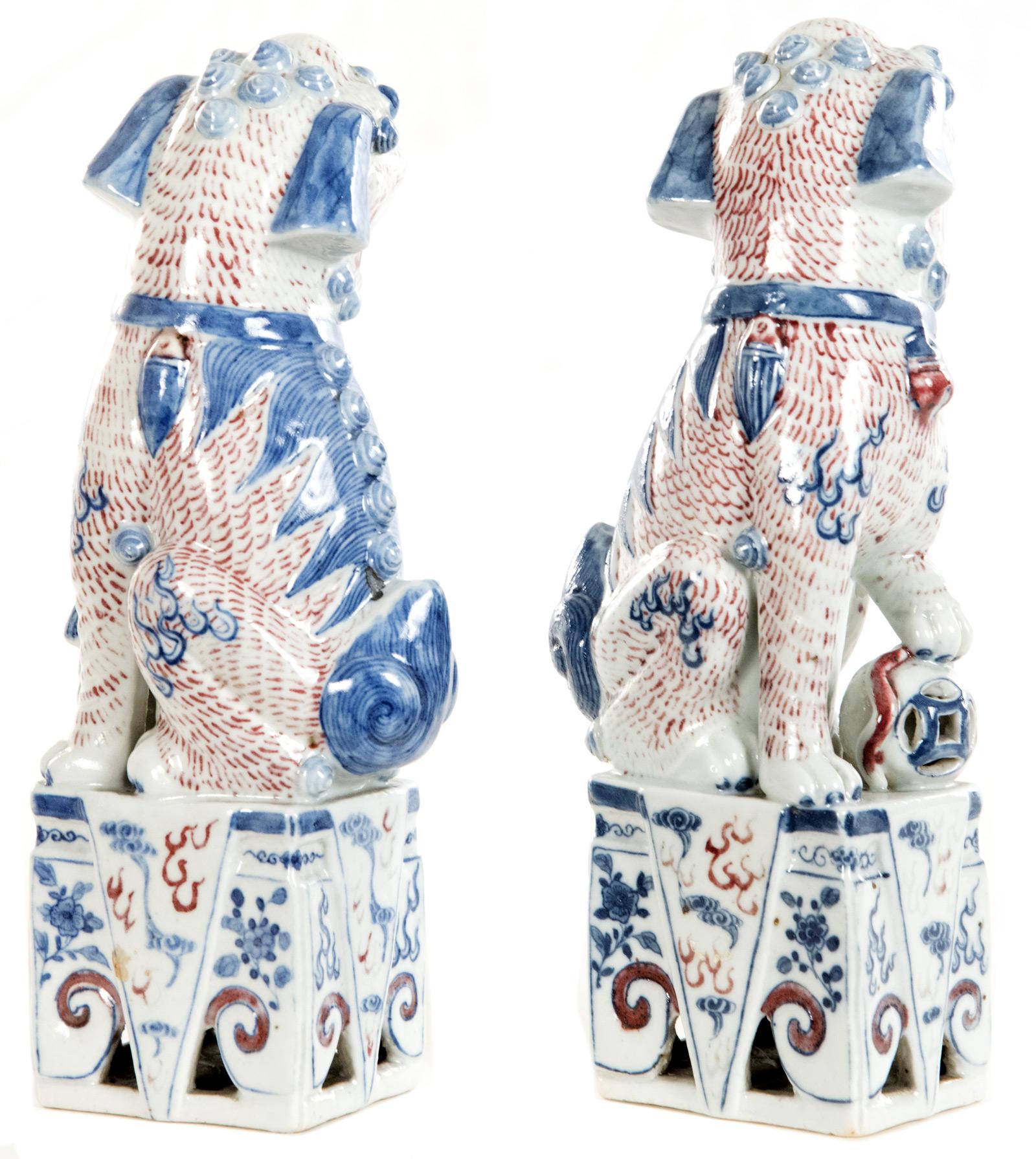 A pair of hand-painted polychrome Chinese foo dogs having a white ground colour decorated with red and blue stylized details, the male with a paw on a ball and the female with a suckling cub, raised on square pierced plinths. Chinese Foo dogs are
