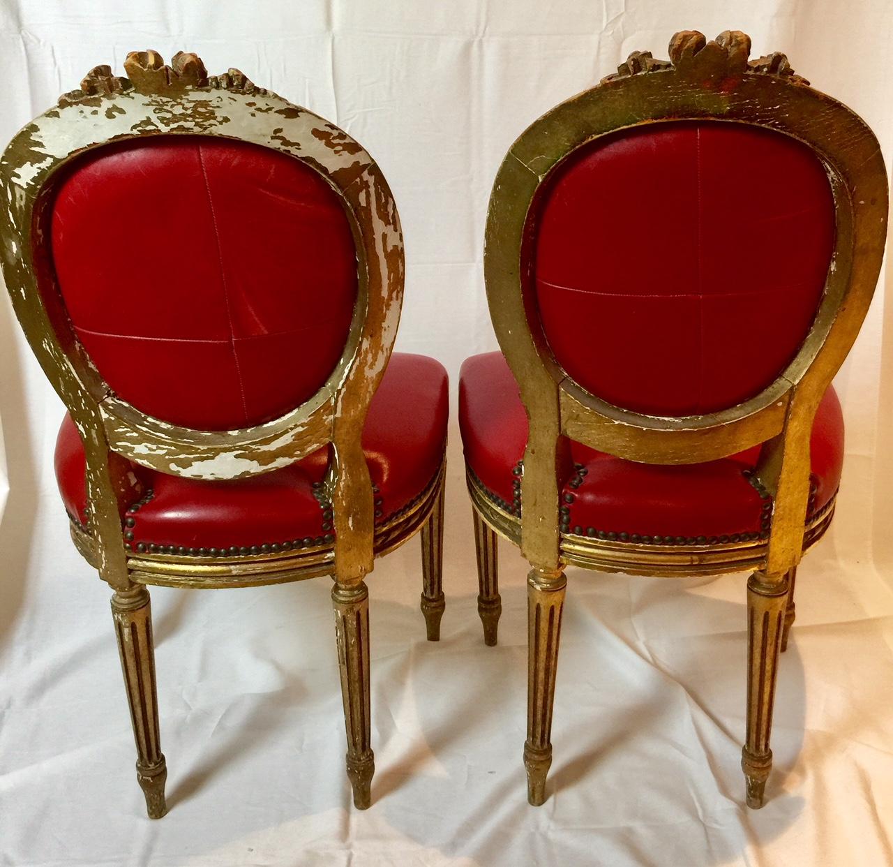 A decorative pair of French giltwood Louis XVI style side accent chairs with carved flower garland and rope detailing, cabriolet medallion or oval back, carved rosettes and fluted (canelure) legs. Red leather upholstery with brass studs, circa 19th