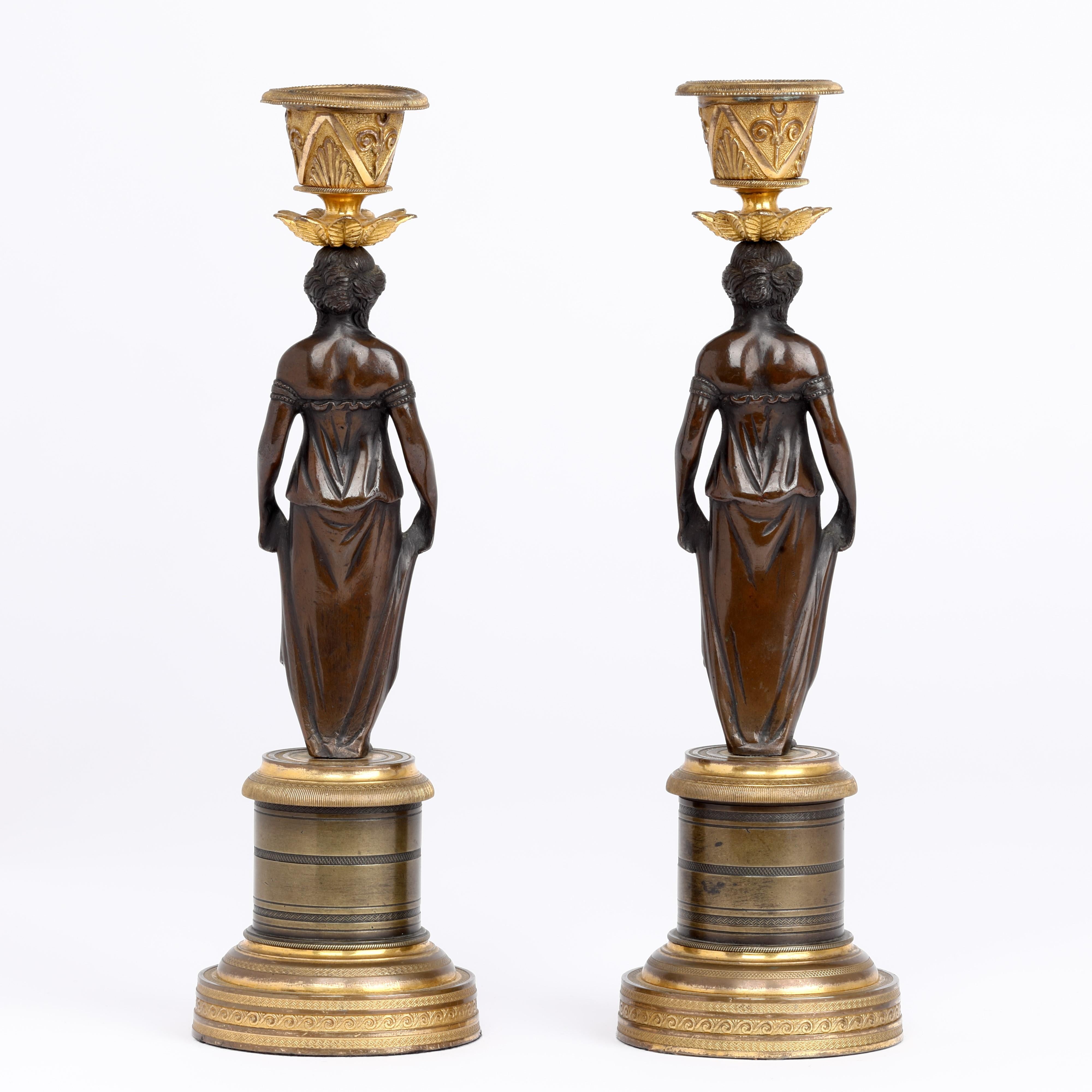 A very fine quality pair of Regency bronze and ormolu candlesticks depicting young maidens with revealed breasts and holding their long dresses above their ancles. Each supports fine ormolu sconces apon their heads with floral drip trays