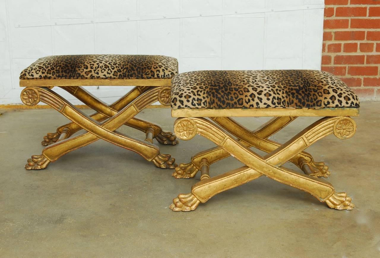 Fabulous pair of Italian Regency carved curule benches or stools featuring a gilt finish and leopard print upholstery. The x-form frames have neoclassical accents and rosettes in the top corners. The stools are supported by lovely lions paw carved