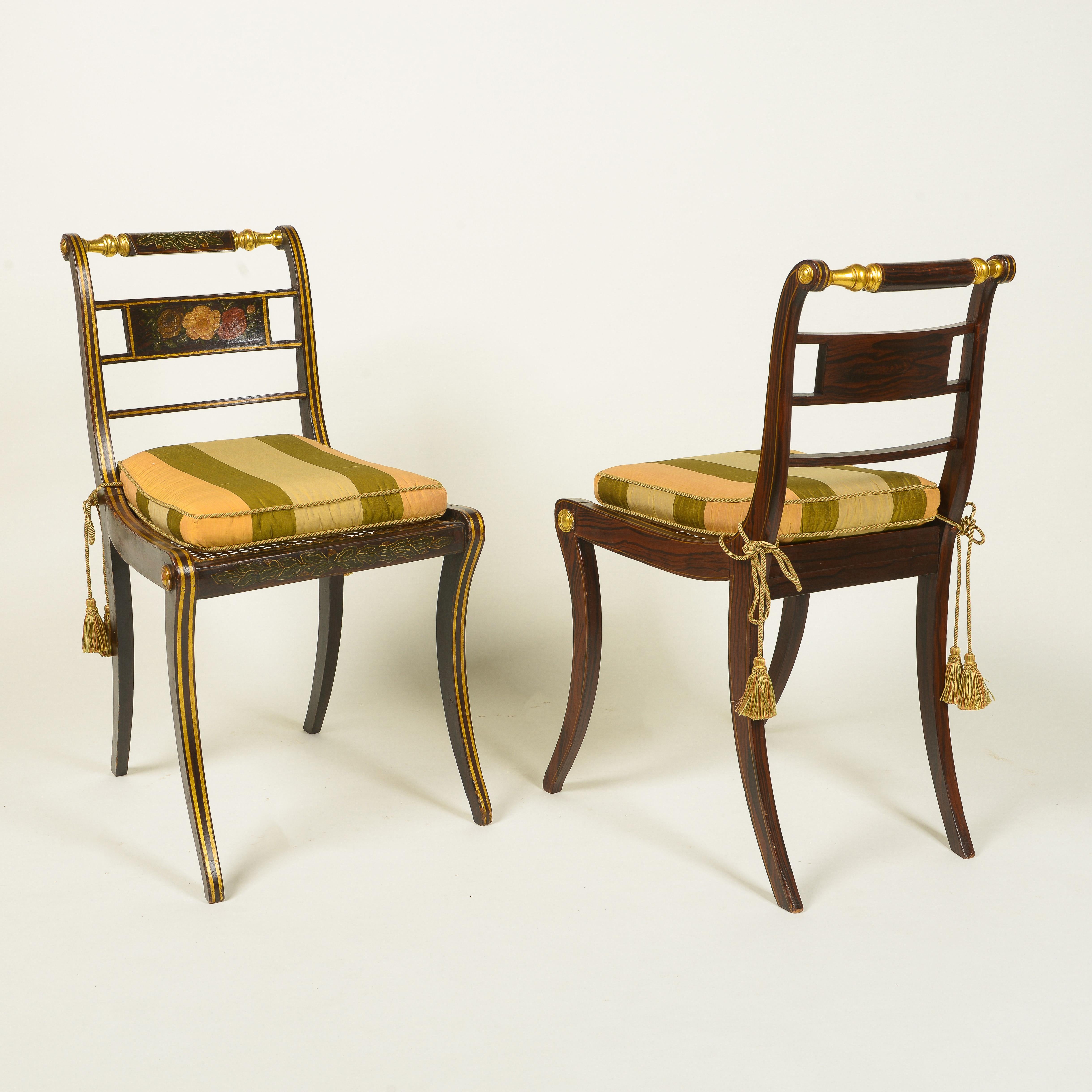 Each back centered by a horizontal splat decorated with different floral specimens; the caned seat striped silk squab cushion in green and pale peach over a foliate-decorated seat rail; on saber legs; with gilt banding throughout.