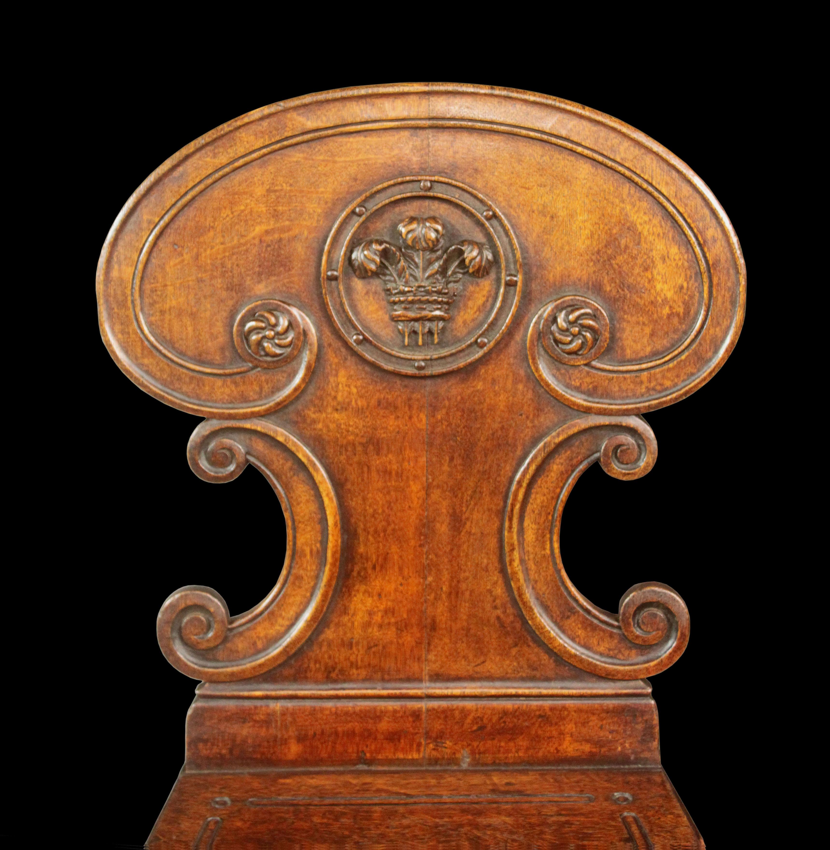 A pair of late Regency oak hall chairs of a good colour and patina; the shaped backs finely carved with C scrolls, patera with spiral carved detail and splendid circular framed Prince of Wales feathers; the seats unusually also with carved details