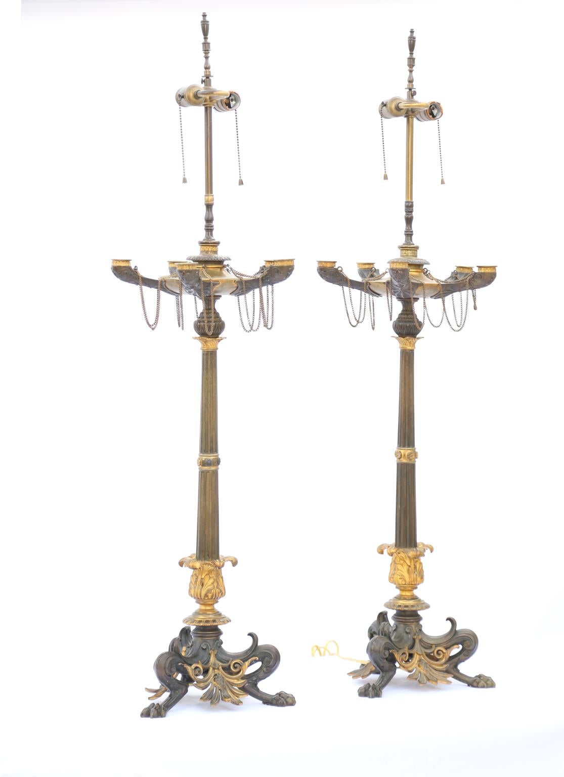 Paire de candélabres à lampes, attribuée à Ferdinand Barbedienne, finement moulée en bronze patiné et doré ; présentant une triple base en forme de pied de biche avec des pattes de pad, décorée de palmettes percées, surmontée d'une colonne cannelée,