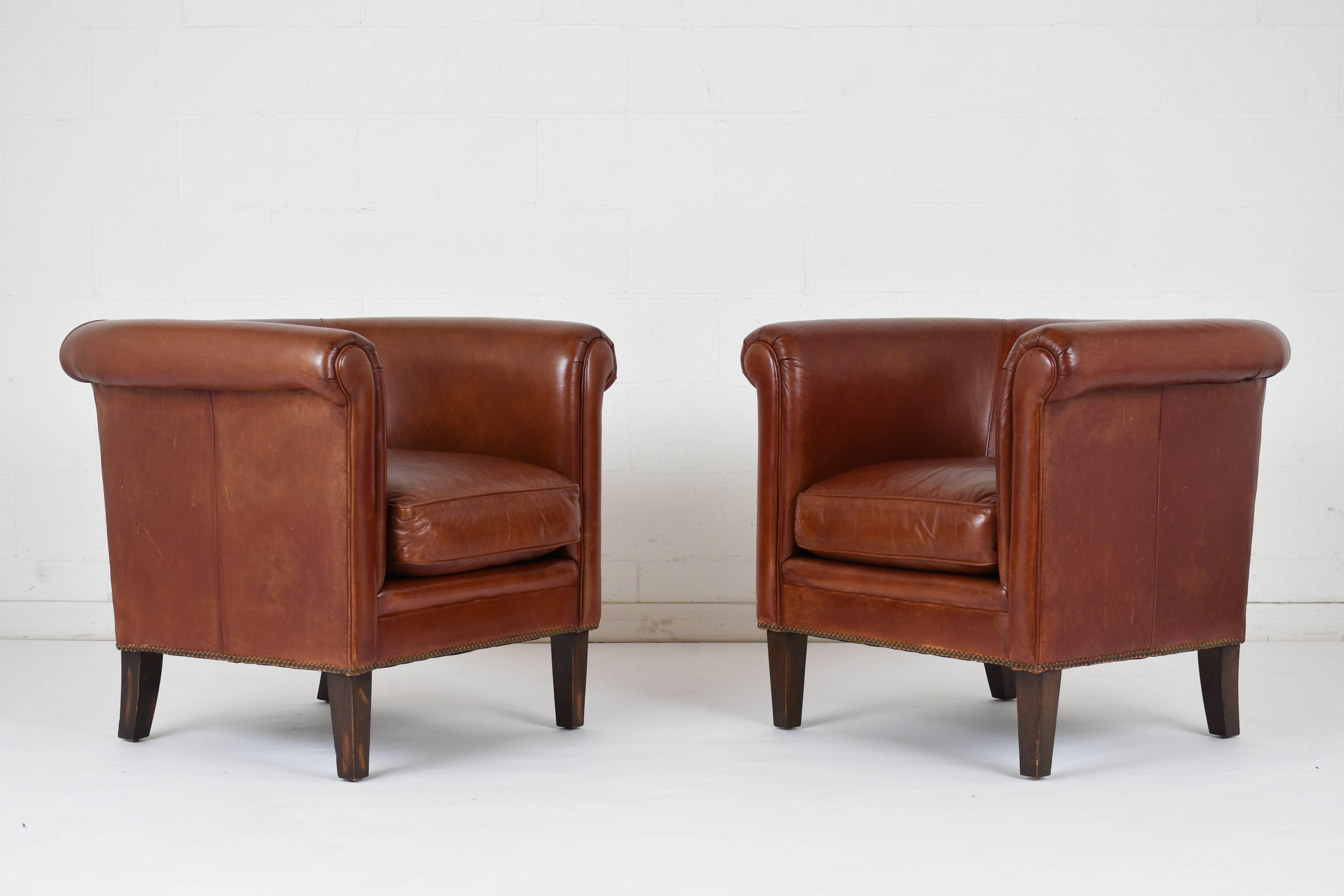 This pair of 1970s Regency-style club chairs are made by Bernhardt and have a traditional profile with a curved back and scroll arms. The seats feature the original cognac color leather upholstery with single piping trim and brass nail head details.
