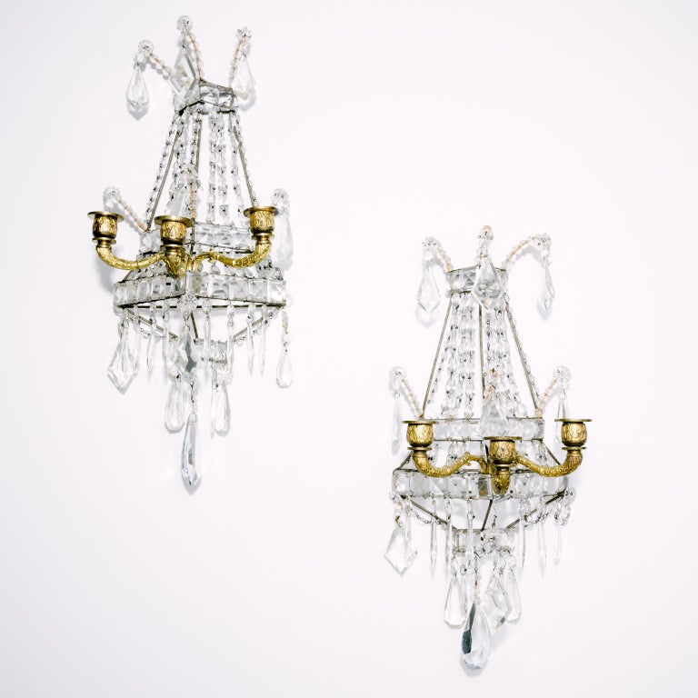A rich and classic Regency style pair of three light wall sconces adorned with faceted crystals and glass decorations of varying uniformed shapes. The candle portion are bronze with gilt metal overlay.