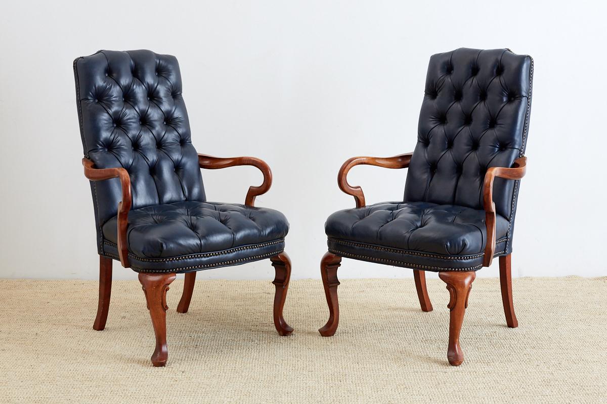Distinctive pair of tufted blue library chairs made in the English regency taste. Features a Chesterfield style tufted navy blue upholstery bordered by brass nail head tacks. The carved frames have crooked arms and small cabriole legs in front with