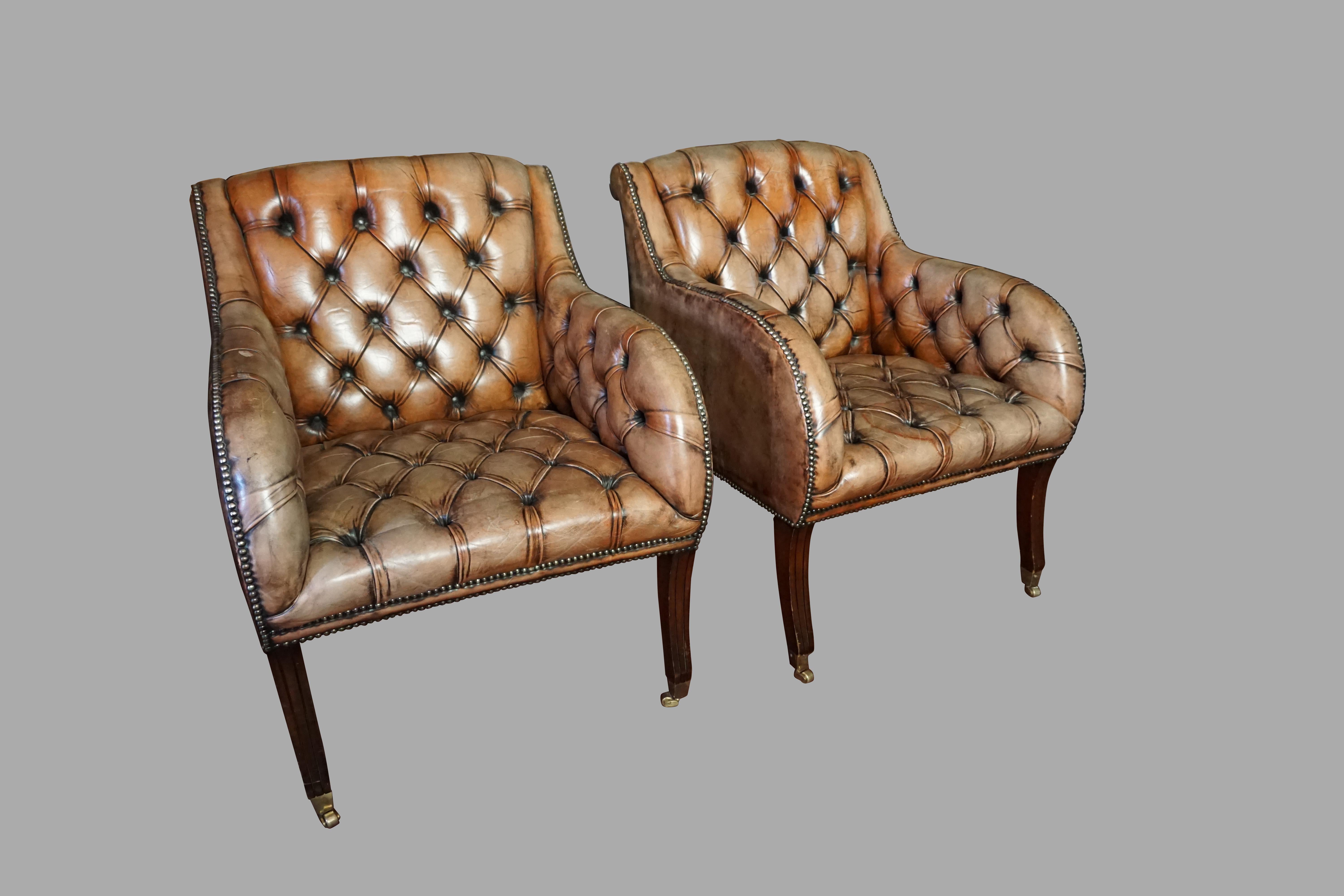A comfortable pair of English tufted leather upholstered armchairs of ample proportions, with rolled leather arms, finished overall in nailhead trim. The chairs rest on molded mahogany saber legs with front brass box casters, 20th century.