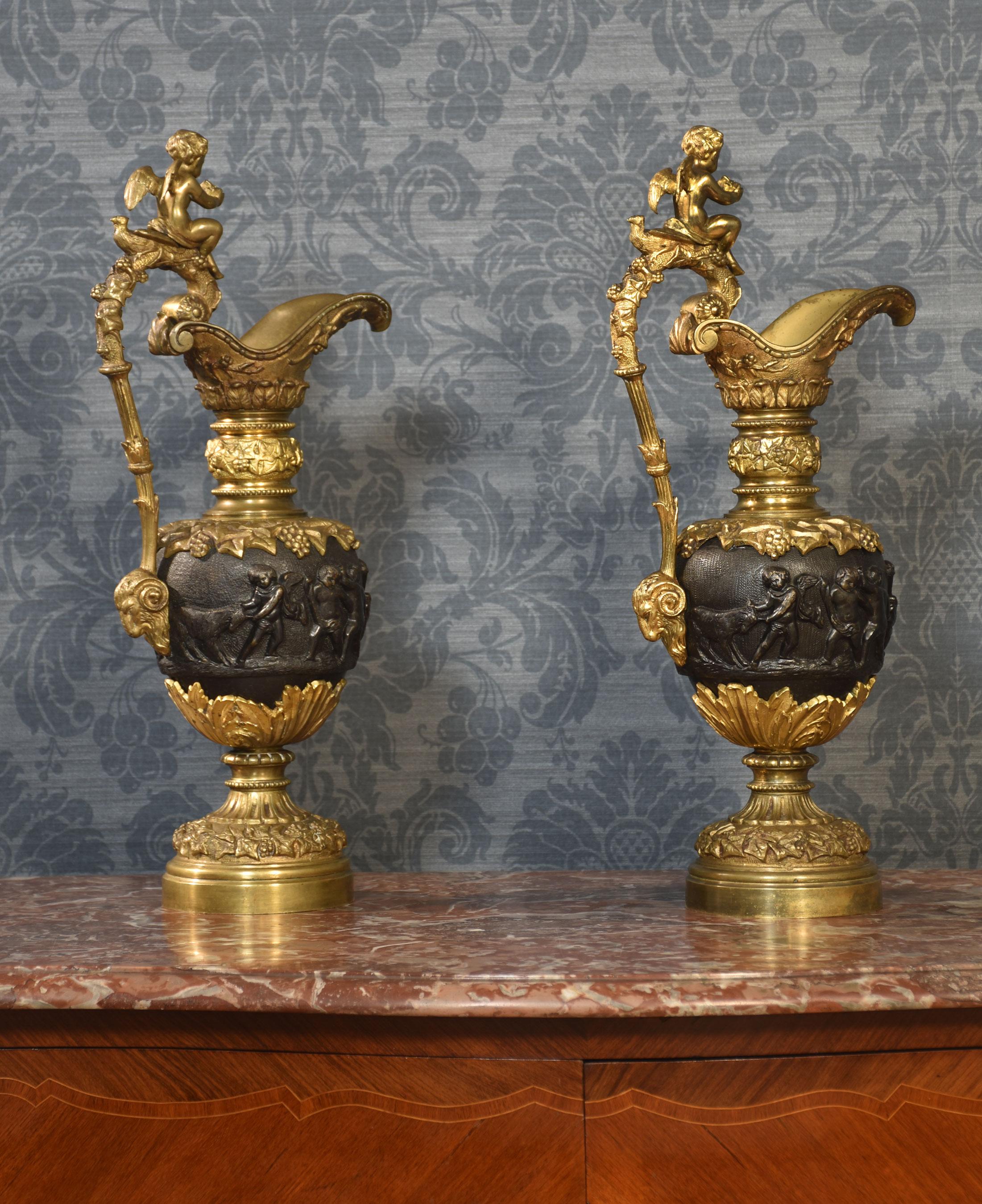 A pair of Renaissance-style French patinated bronze ewers cast overall with trailing grape vines on a punched ground, the handles with cherub mounts issuing from the rims descending to grotesque masks at the bodies, the bodies cast with putti