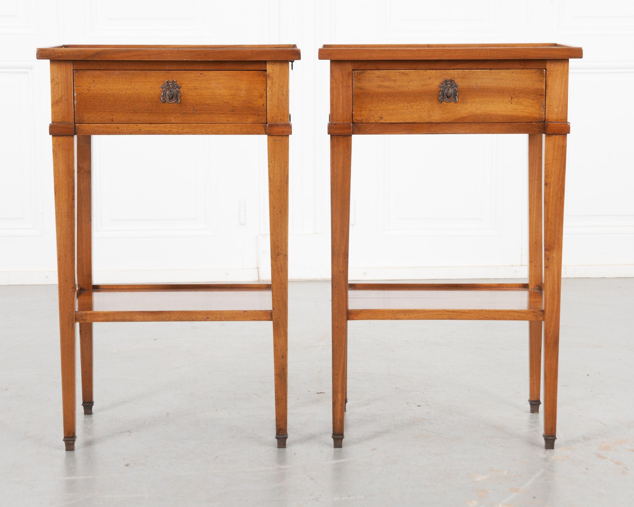 A charming pair of light fruitwood side tables recently constructed in France. A small lip is featured around the top. Each apron houses a single drawer and drink slide. Laurel and ribbon eschutecons grace the front of each drawer; both contain a