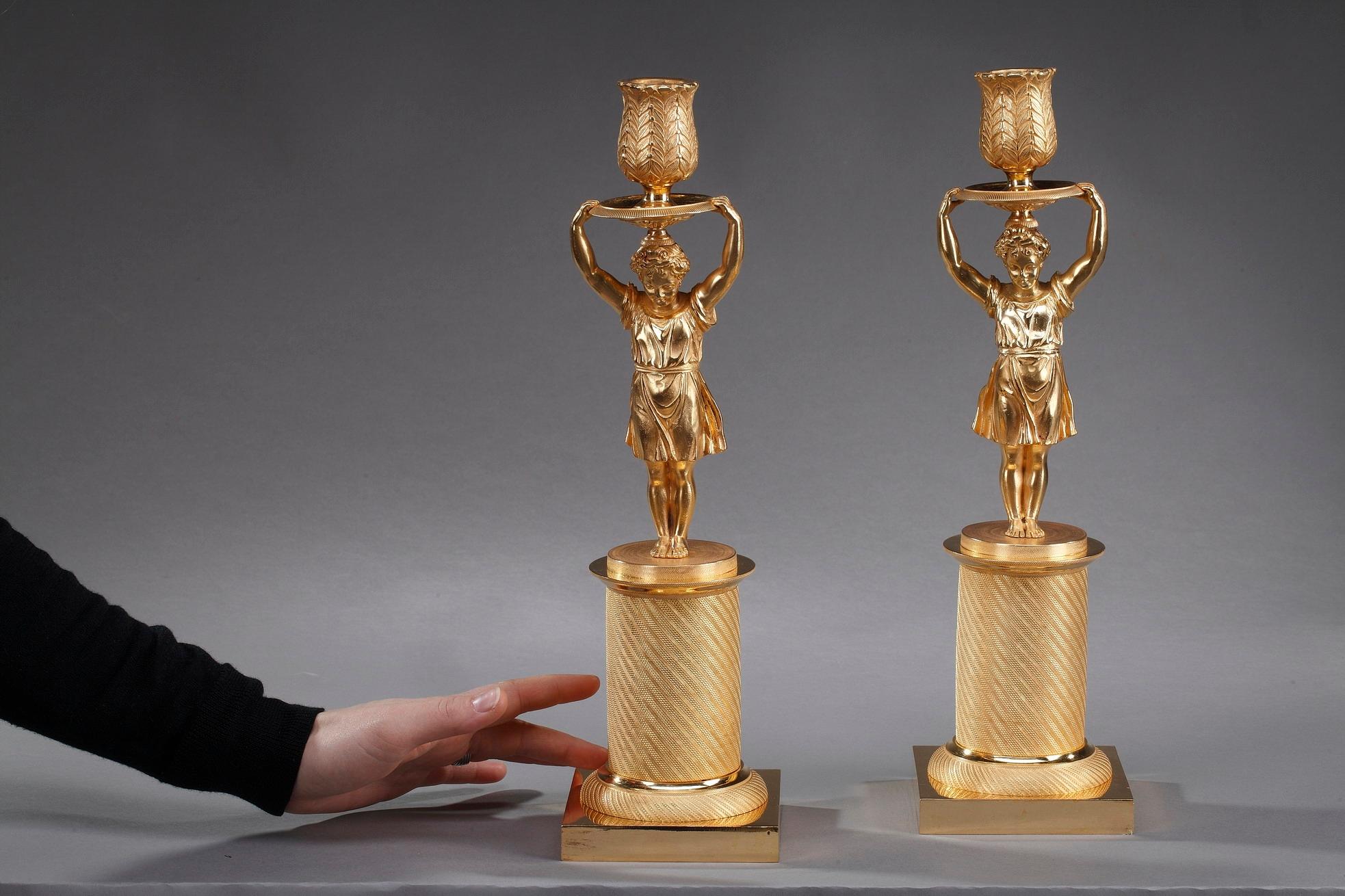 Pair of small restauration candlesticks crafted of gilt bronze featuring a girl standing up on a circular pedestal chiseled with twisted patterns, above a square plinth. She wears a floting dress and holds over her head the socket highlighted with
