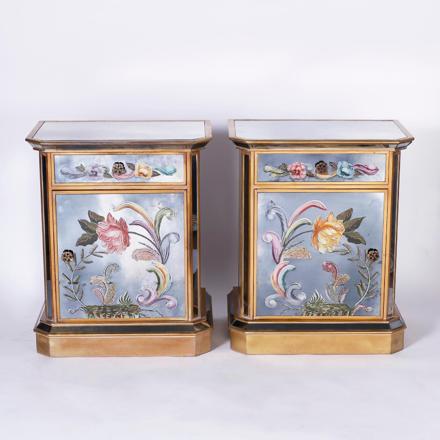 Pair of mirrored night stands or end tables with a Classic form and reverse painted or églomisé panels decorated with dramatic floral designs. Featuring slide out shelves, old world style painted trim, and floral brass hardware.