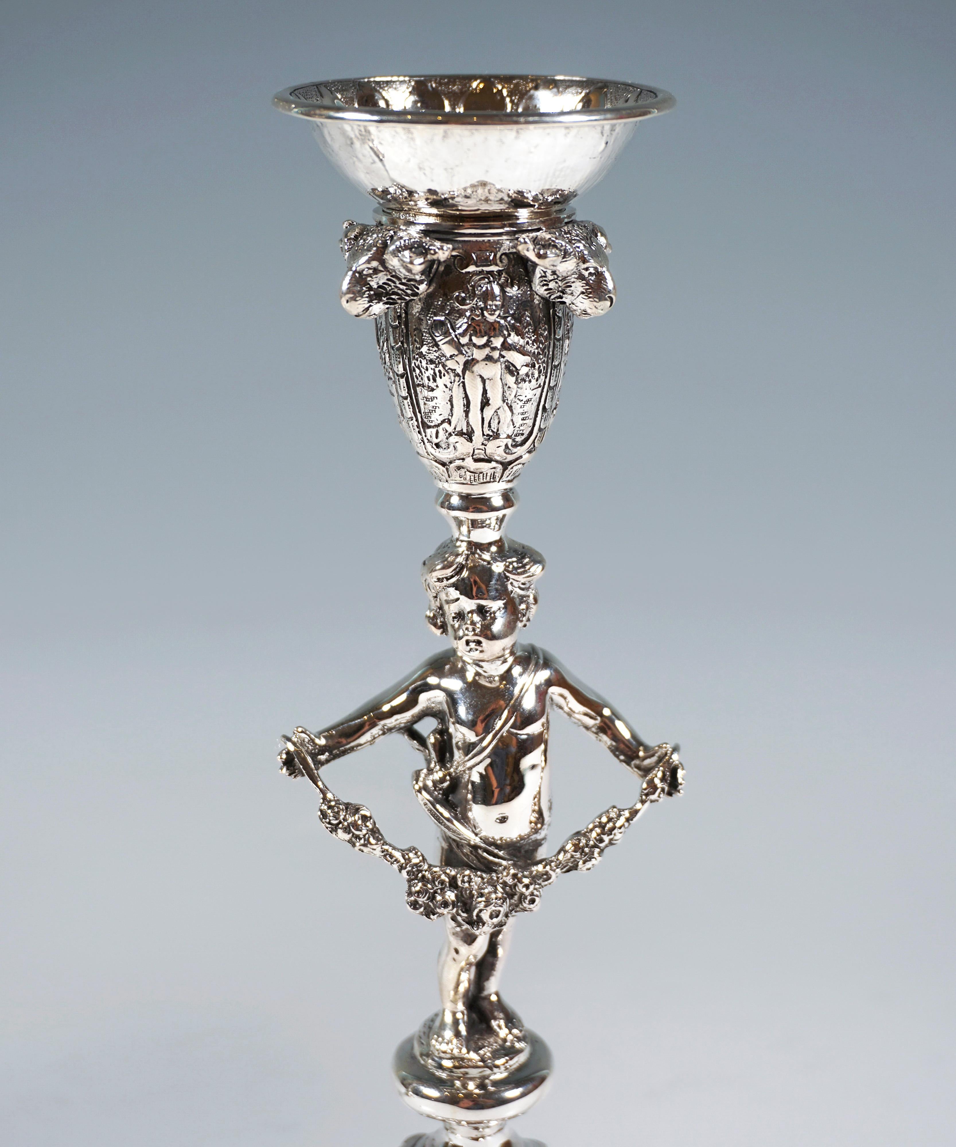 Pair of richly decorated candlesticks on a high base, bulging body, with a fully sculpted putto carrying a garland in place of the shaft, balancing the vase-shaped spout with sculpted rams' heads on its head.
Rich floral decoration, as well as