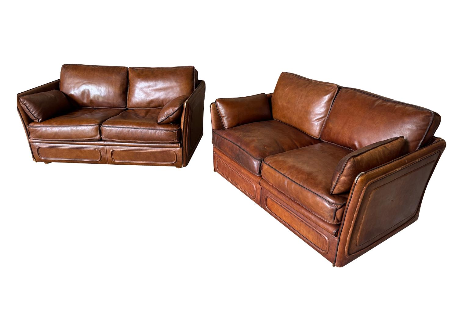 A very chic pair of mid-century Roche Bobois Sofas from France. Handsomely crafted in vicuna toned leather with with stitched edges, metal corner details and very comfortable cushion seats. One sofa measures 30 1/2