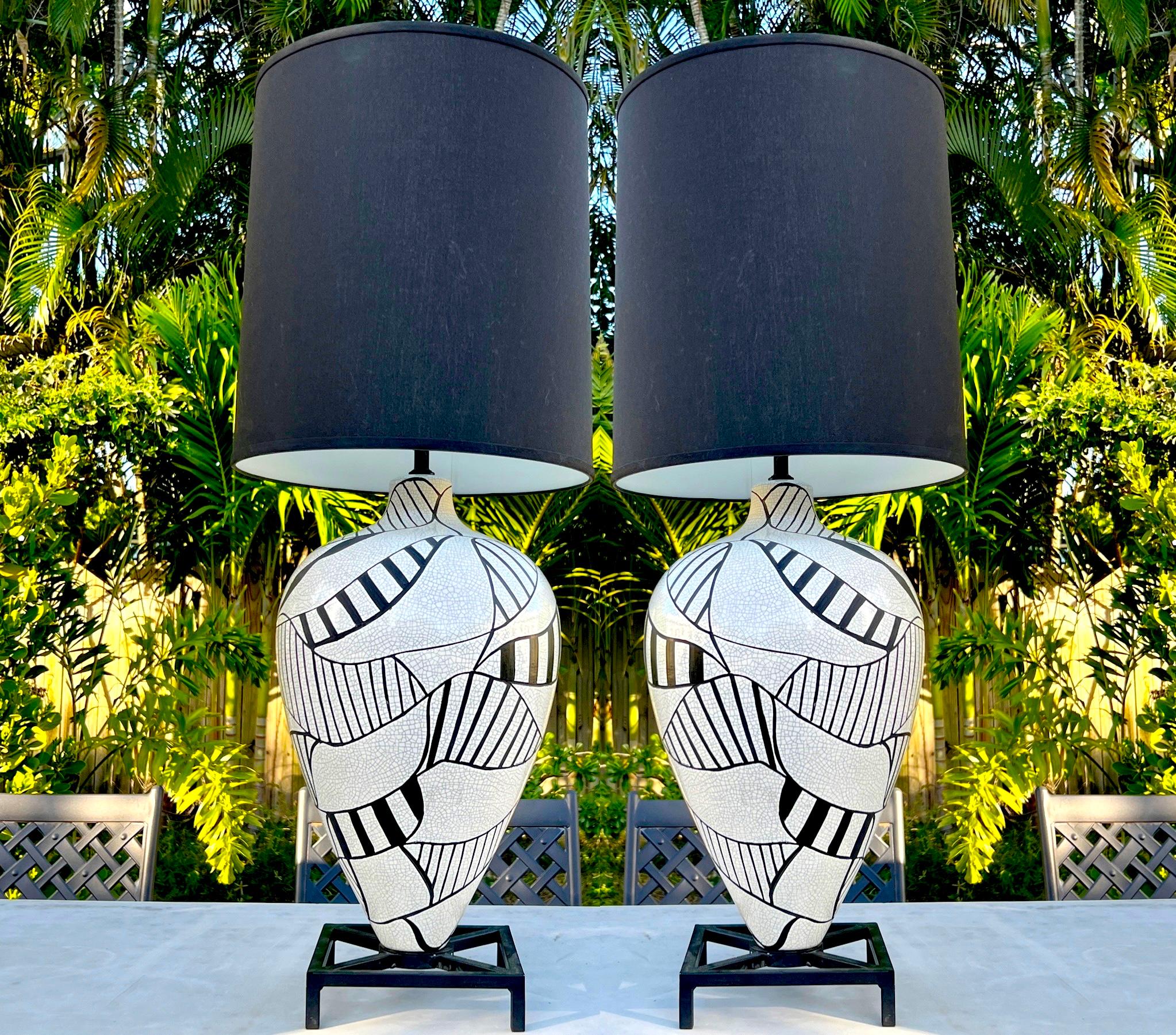 Pair of ceramic pottery lamps with large-scale urn forms featuring a white crackle glaze finish with hand-painted geometric designs in black enamel. The vintage lamps sit on footed iron bases in black. The lamps have a mid-century modern design