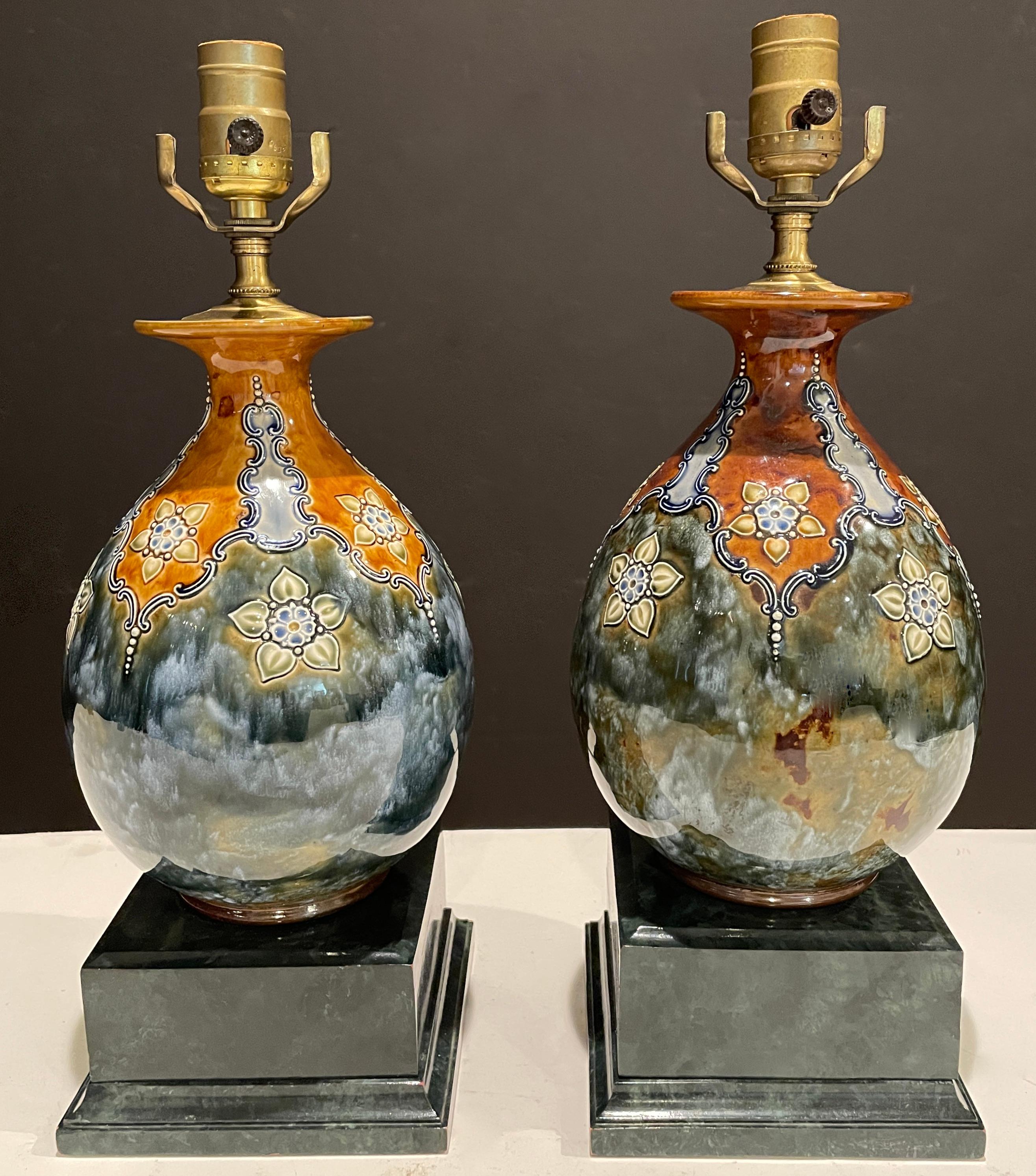 Pair of Royal Doulton Art Nouveau stoneware vases mounted as lamps. Exterior glazed in a blue grey ground with raised stylized floral decoration in greens and browns. Raised on marbleized square wood base.
24