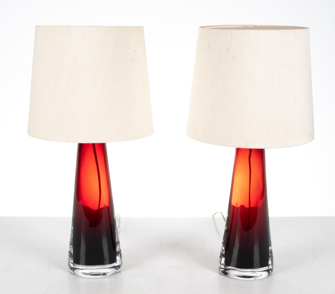 This pair of jewel-like lamps features heavy bottle-form bases of cased clear-over-ruby red glass, with chrome socket mounts and cream raw silk shades. This form was designed by Carl Fagerlund and produced by Orrefors in the 1950's and 1960's; each