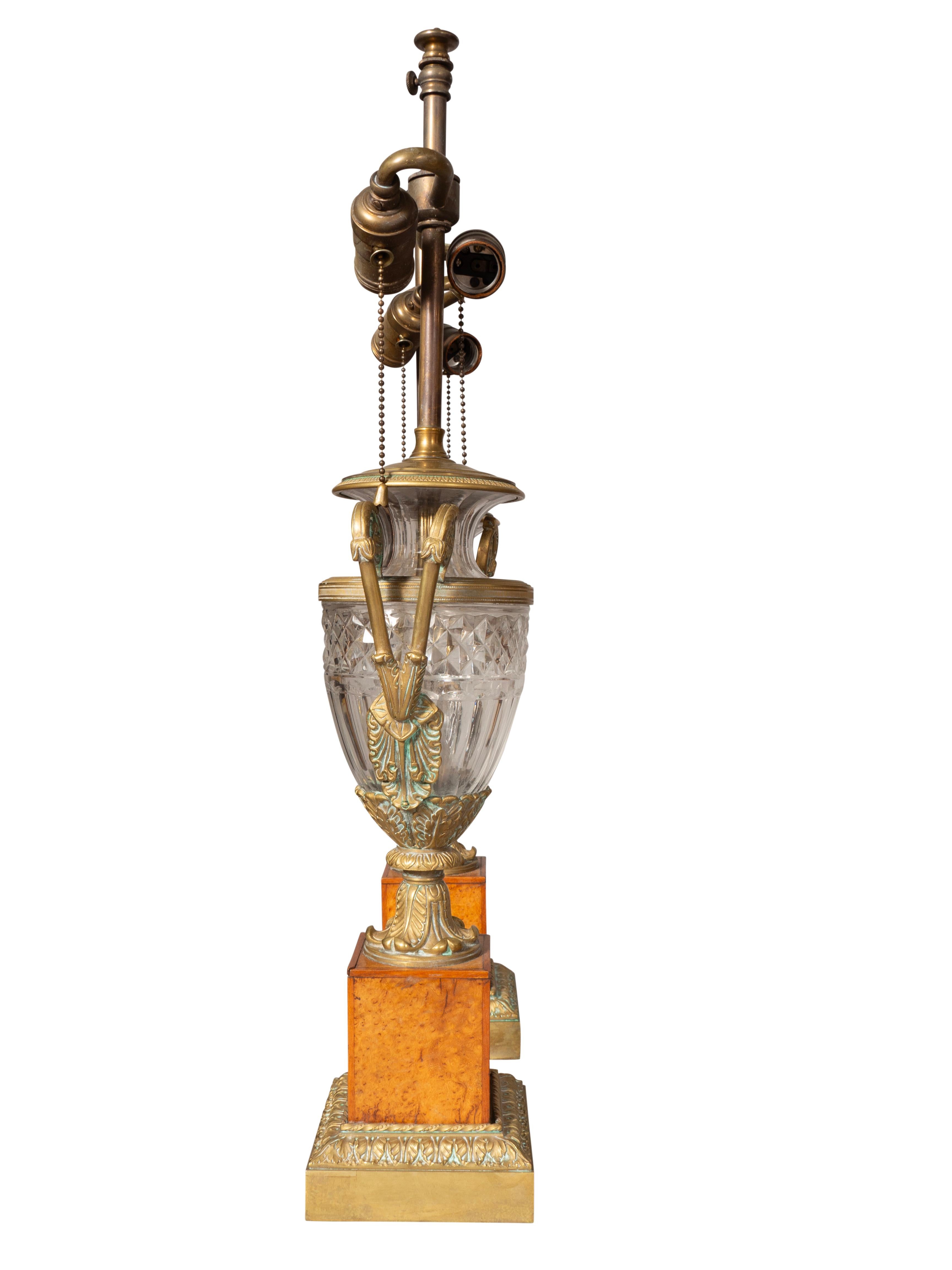 Each with two lights. Cut glass urn form vases with bronze handles seated on a burl wood and bronze mounted plinth. 