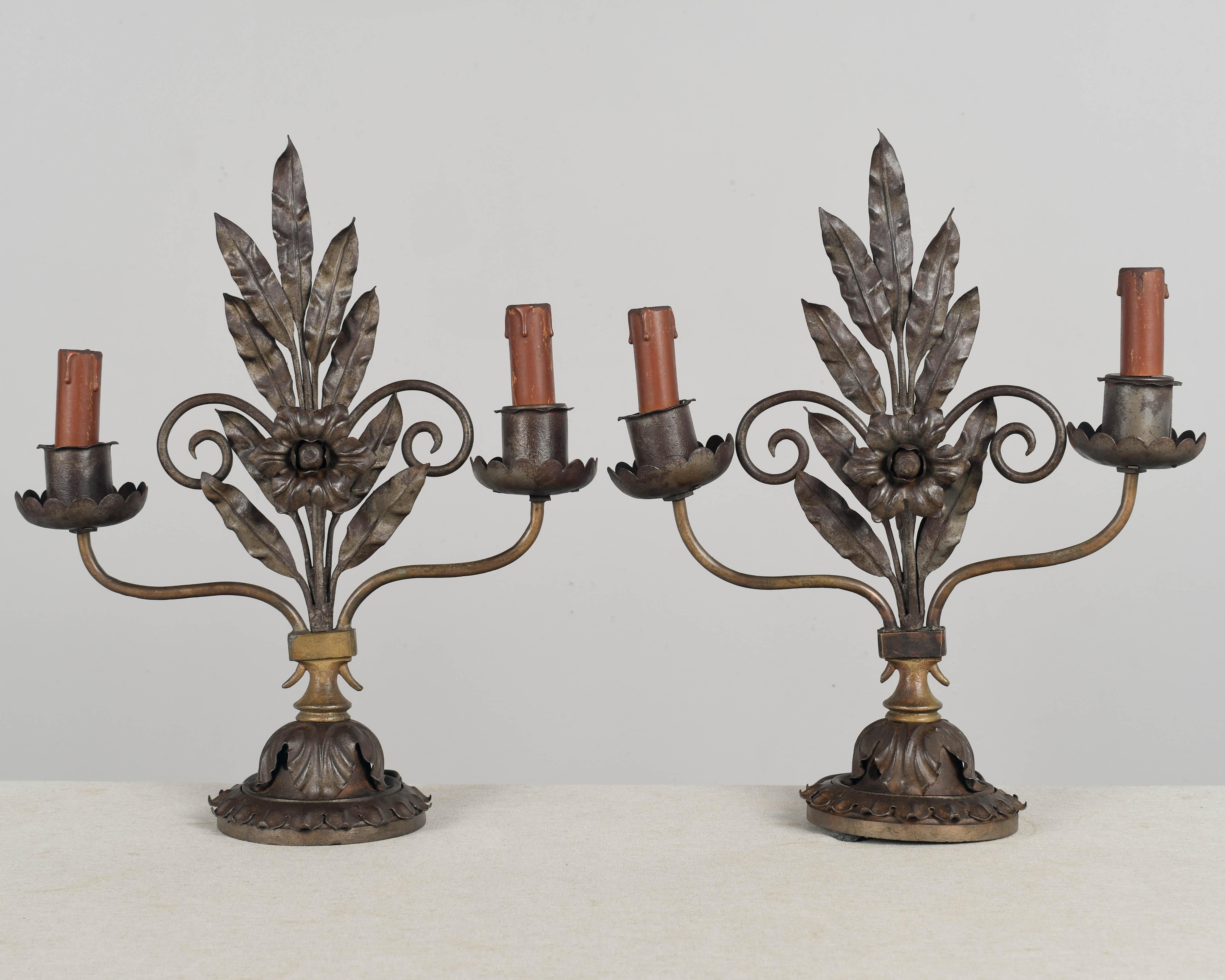 A pair of rustic cast iron and patinated metal two-light candelabra with flower leaves and scroll forms. Original candle covers. Rewired with new sockets and cord. Rustic condition with some rusting. A little uneven, one is a little crooked and